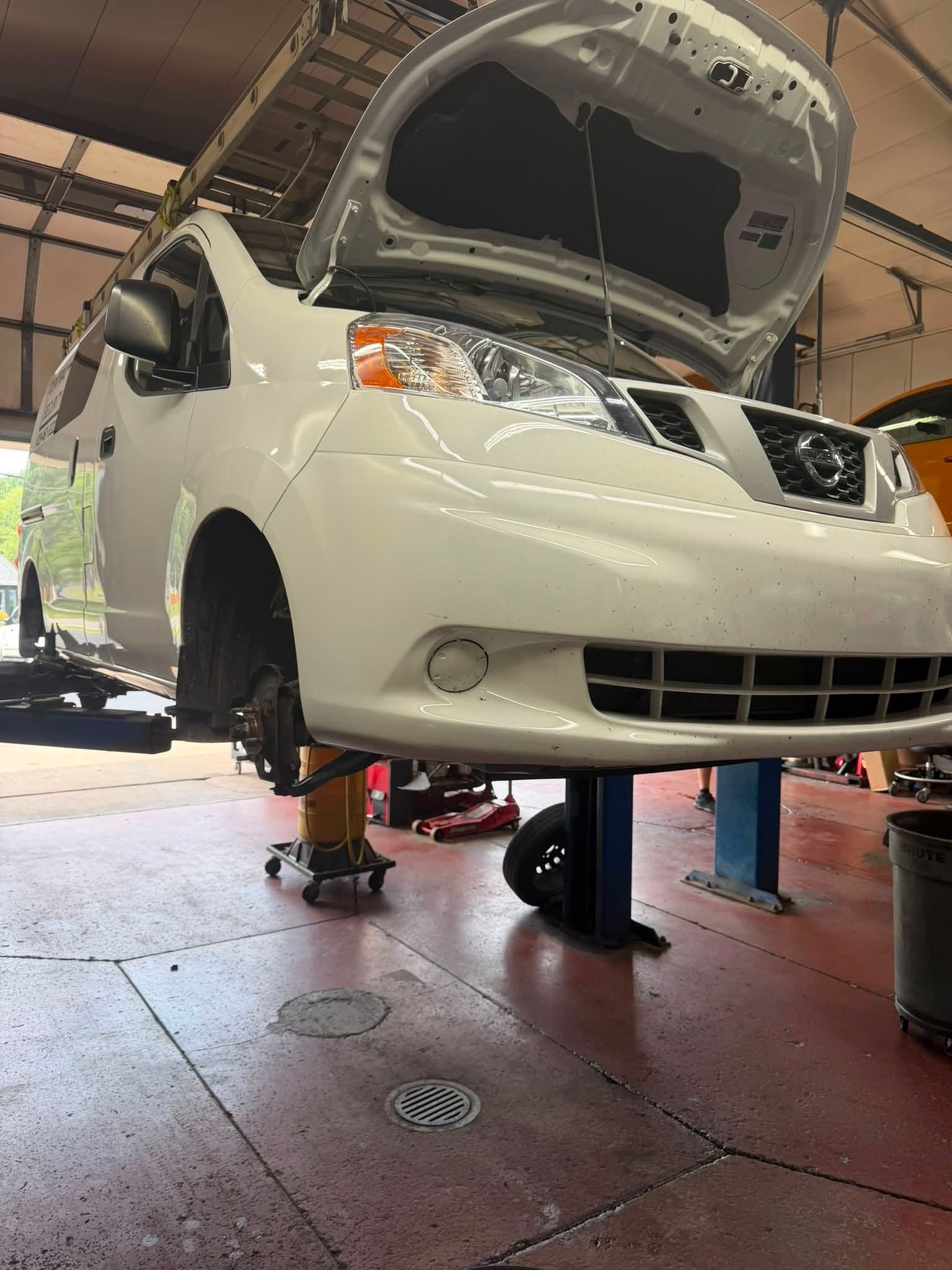 White car on a lift in a garage with its hood open and front wheel removed.