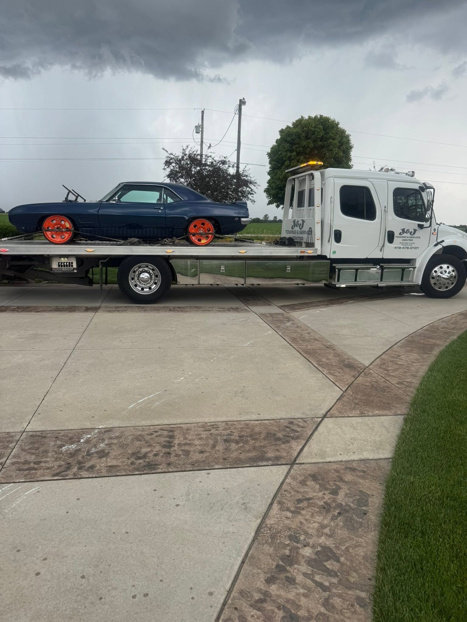 Tow truck carrying a black car with orange rims on a driveway under cloudy skies
