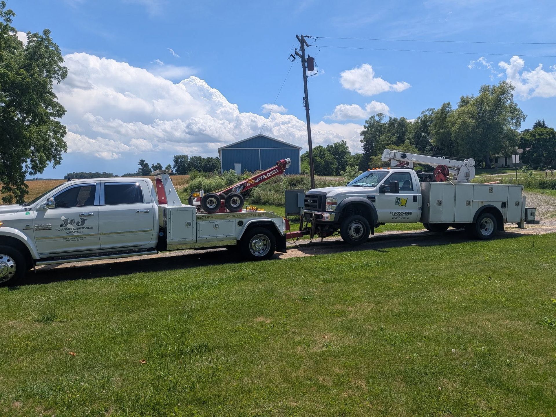 Two white tow trucks parked on grass under a blue sky, with a building and trees in the background