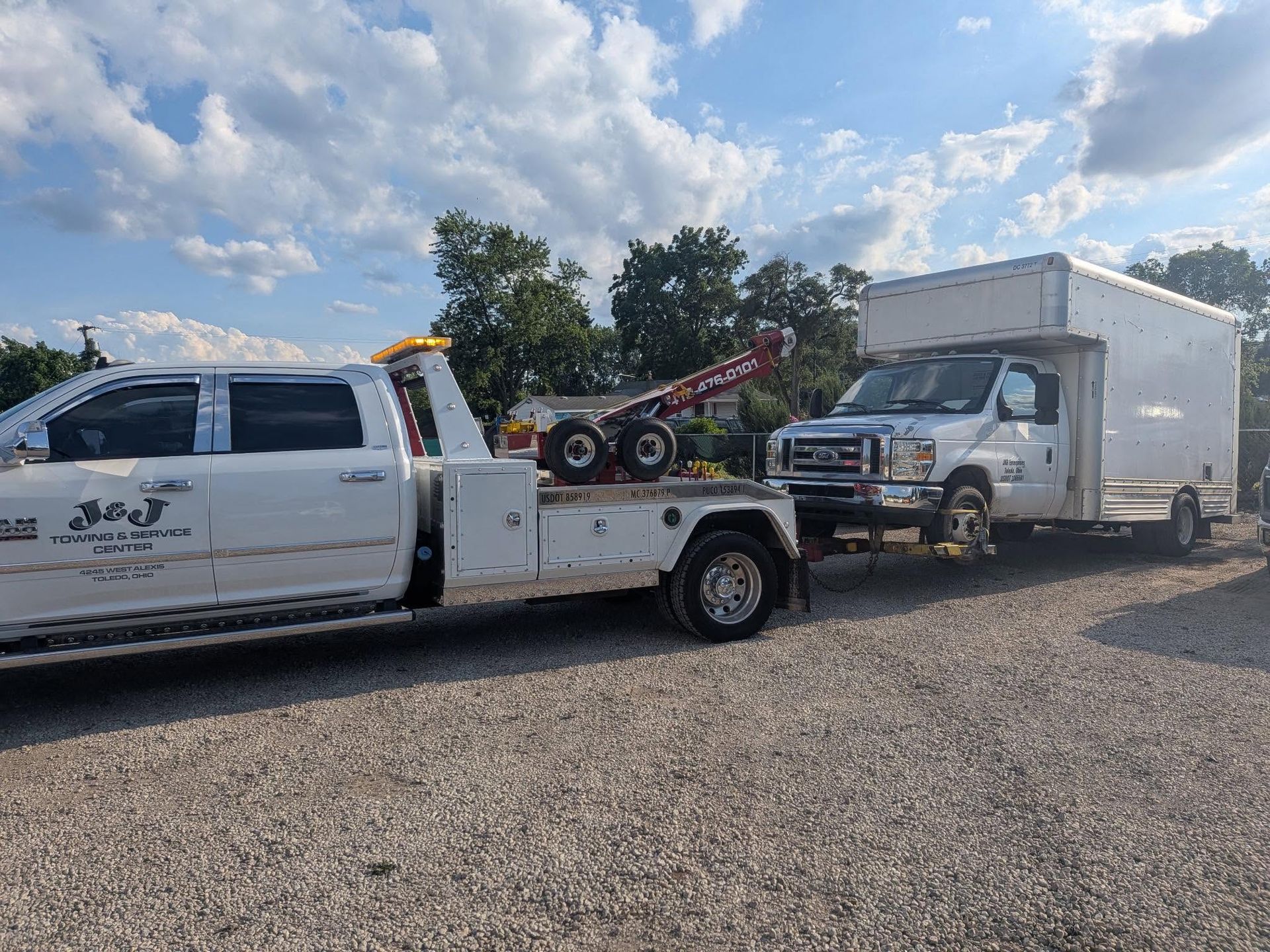 White tow truck hauling a box truck on a gravel lot under a cloudy sky