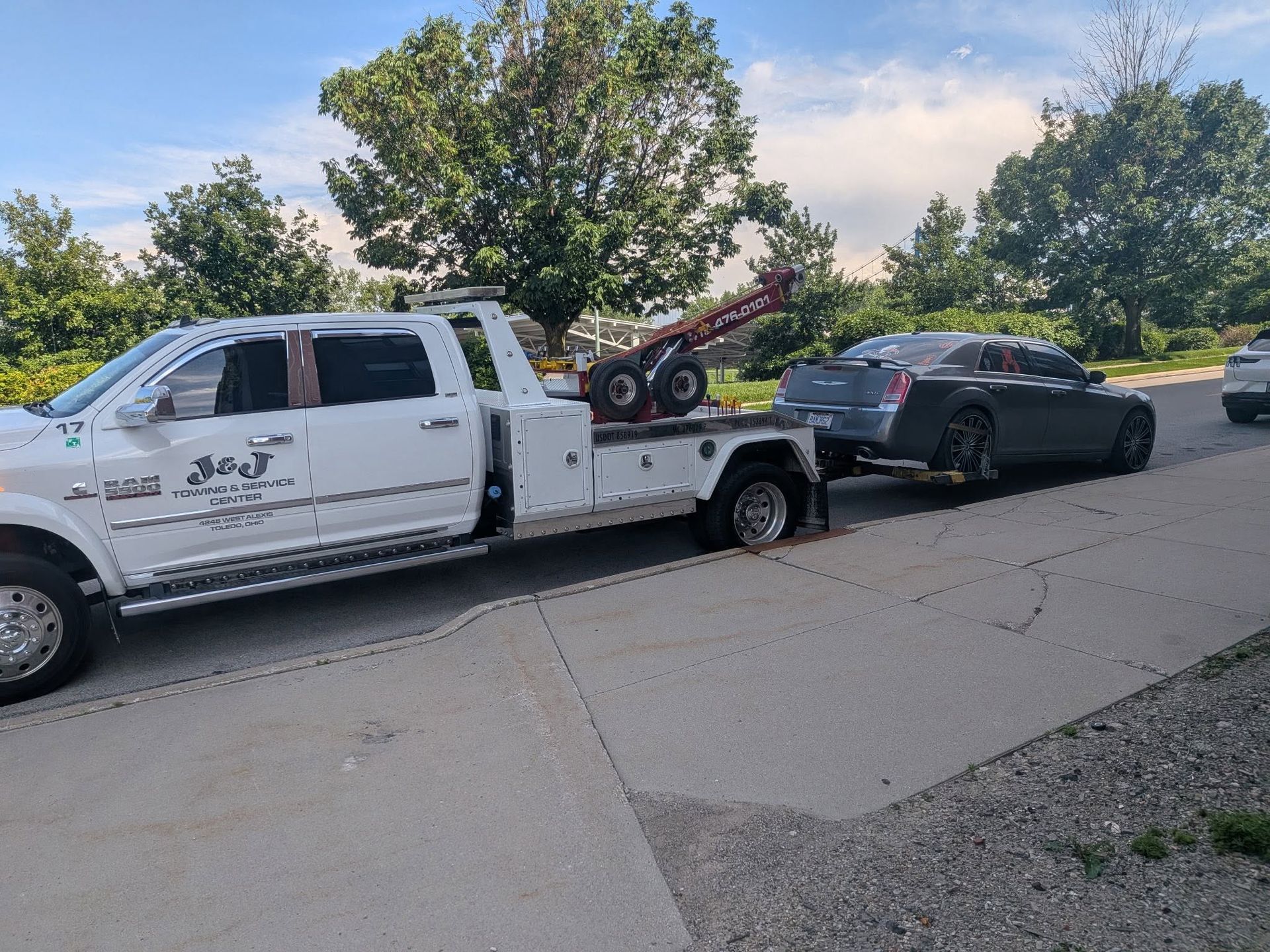 White tow truck hauling a black sedan on a residential street beside trees and parked cars