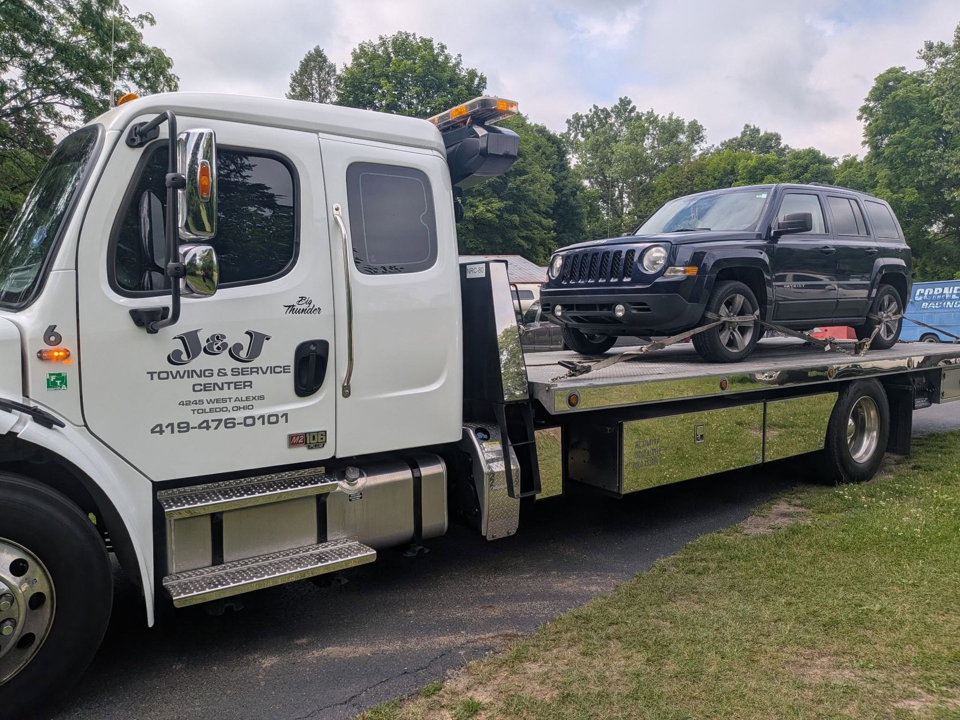 White tow truck carrying a black SUV on a flatbed beside a grassy roadside under cloudy skies