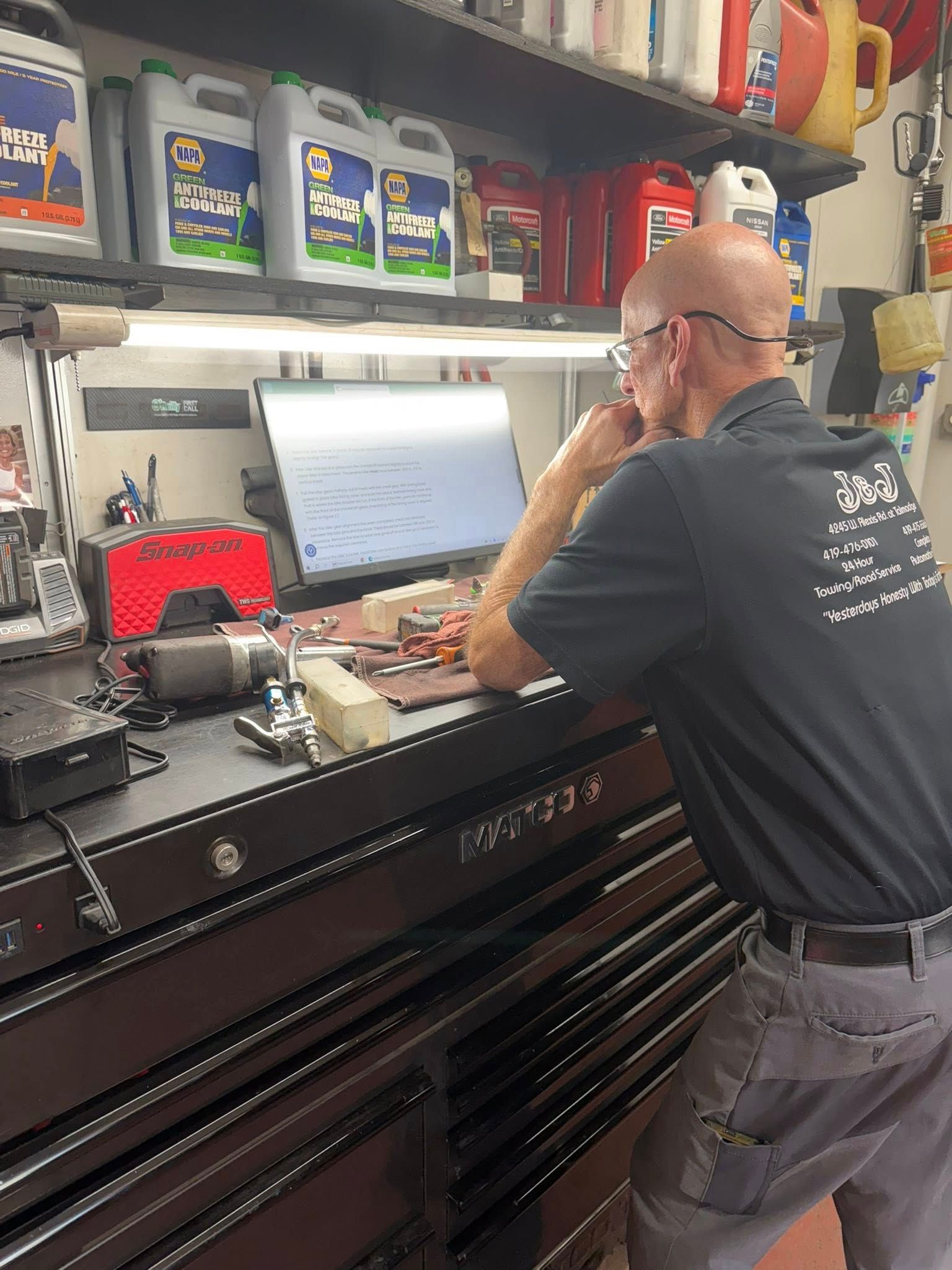 Mechanic using a laptop at a cluttered workbench in a garage, with shelves of supplies behind him
