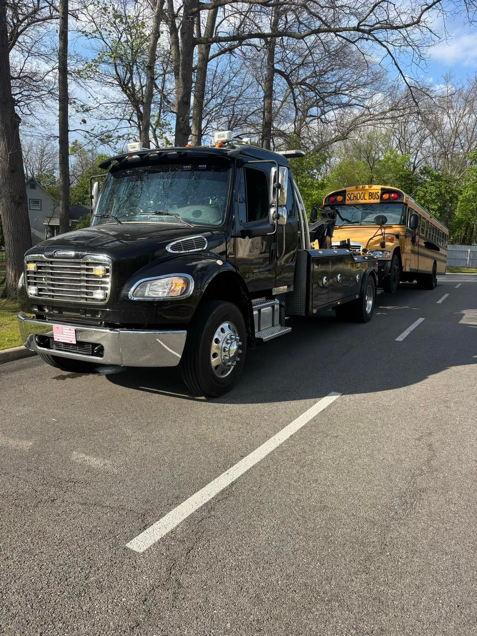 Black tow truck hauling a yellow car on a road lined with trees, sunny day