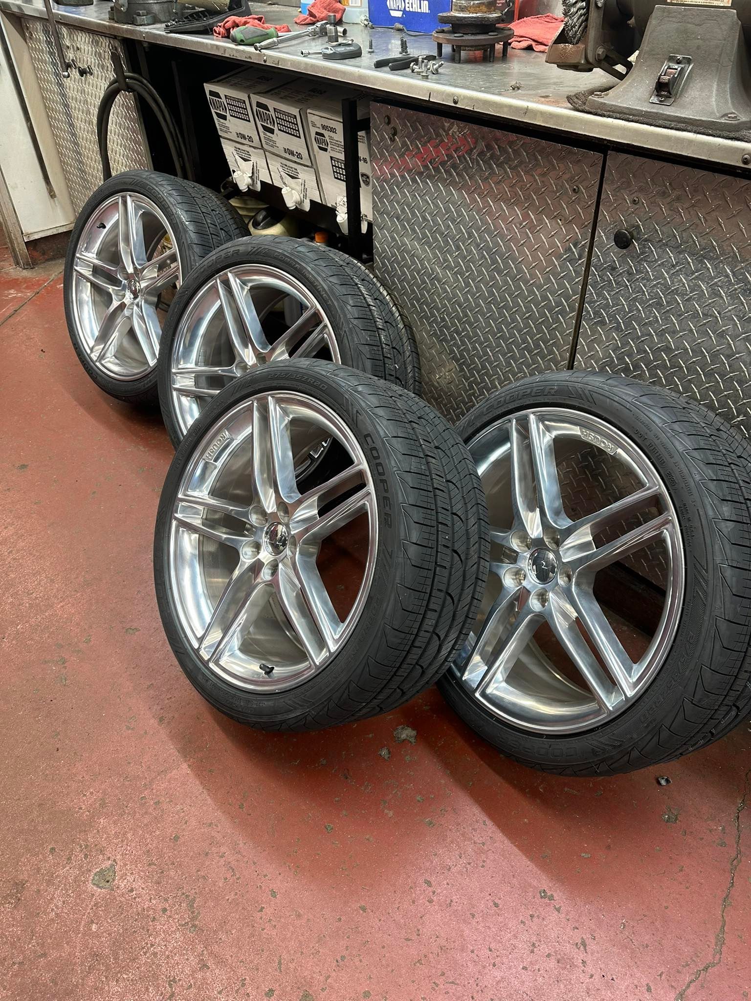Four car tires with silver rims leaning against a metal workbench in a garage