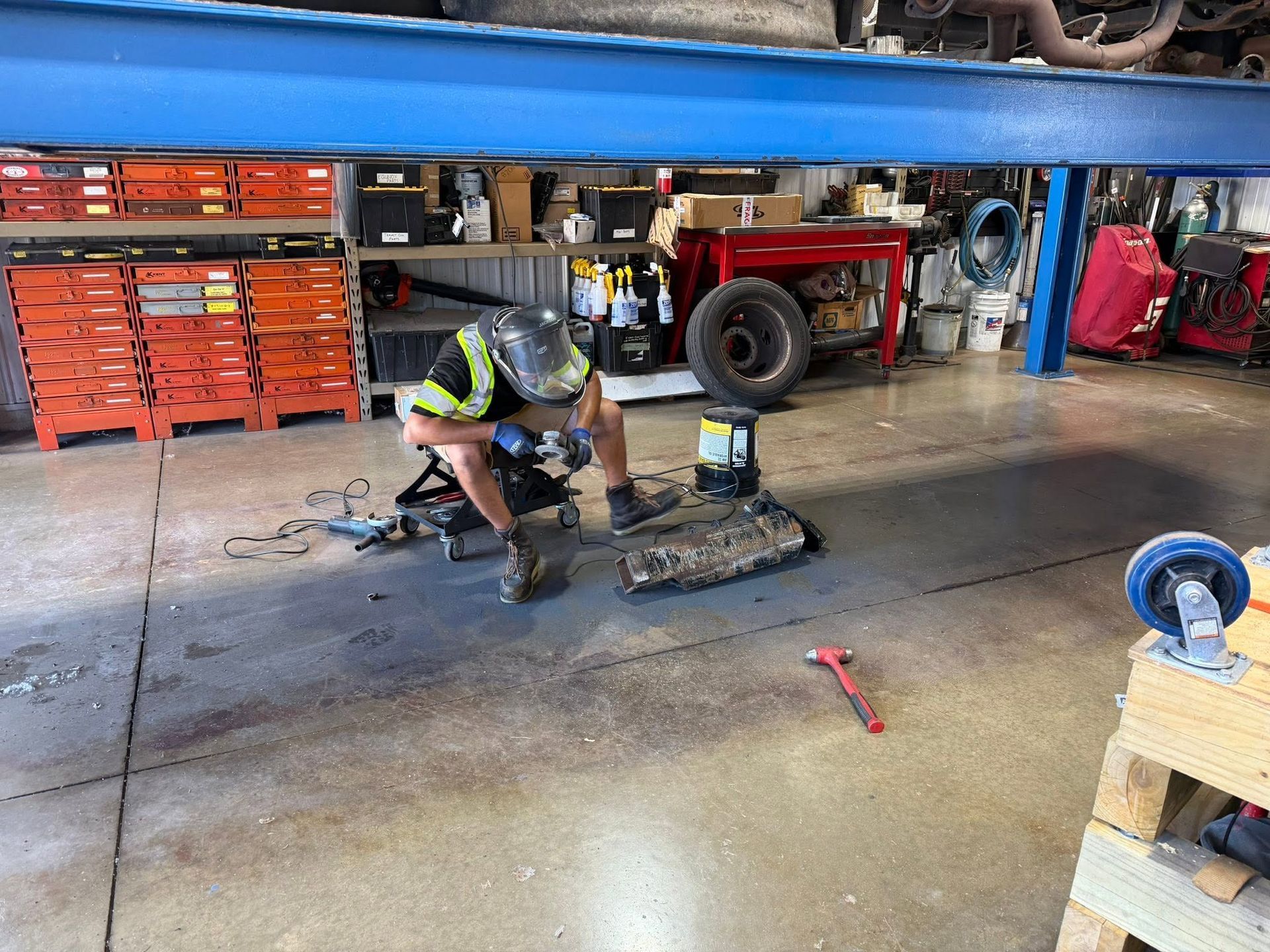 Mechanic kneeling under a vehicle, working on parts in a garage with tools and equipment.