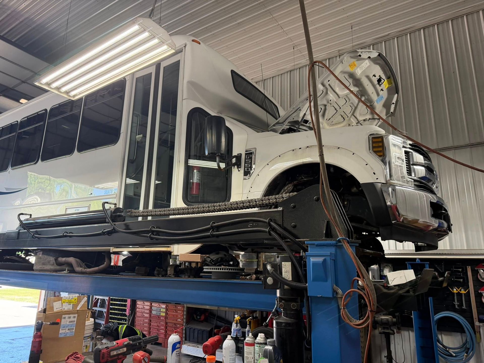 White school bus on a lift in an auto repair shop, with the front end removed and engine exposed.