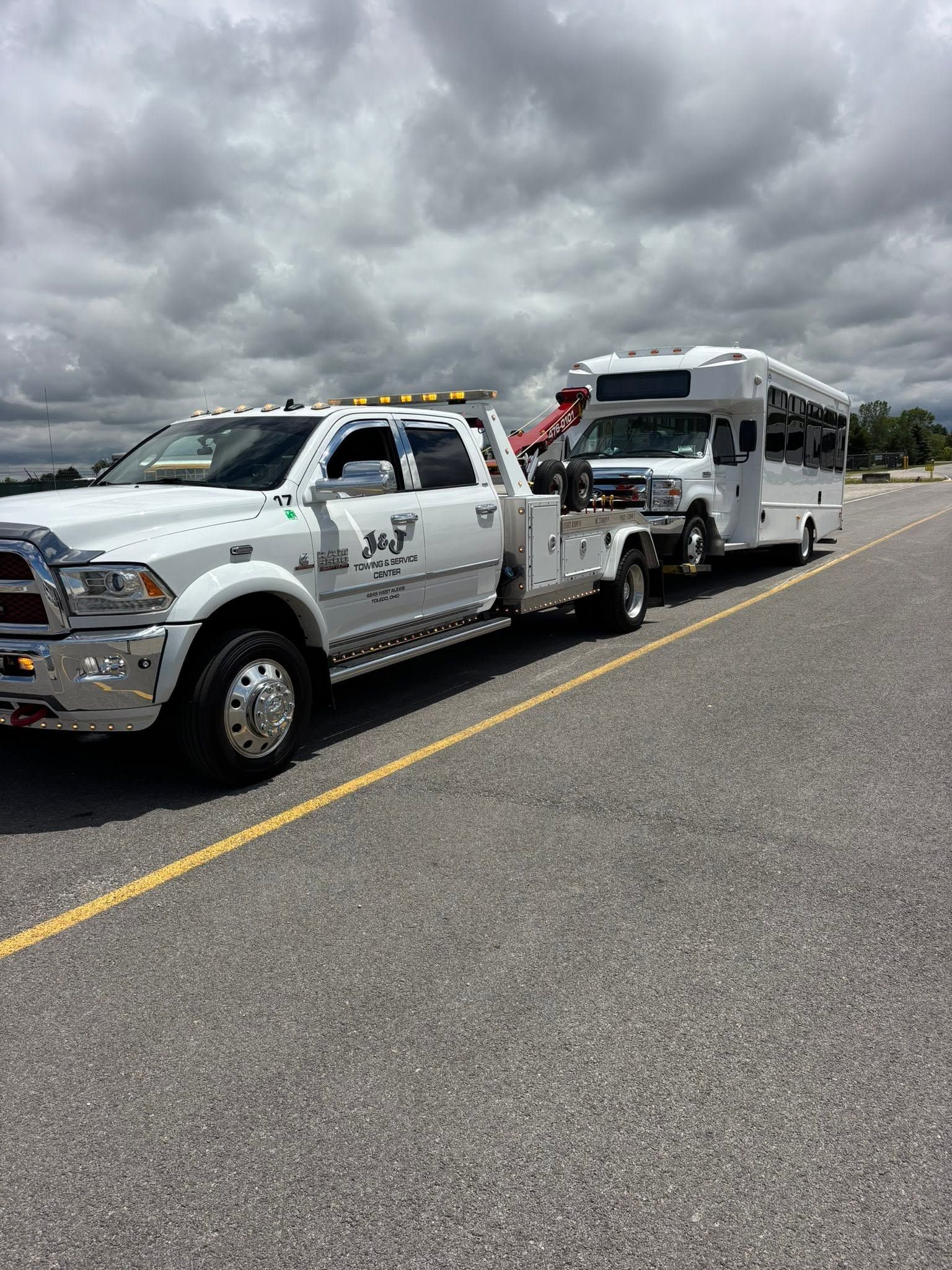 White tow truck pulling a white coach bus on a cloudy roadside with another truck behind them