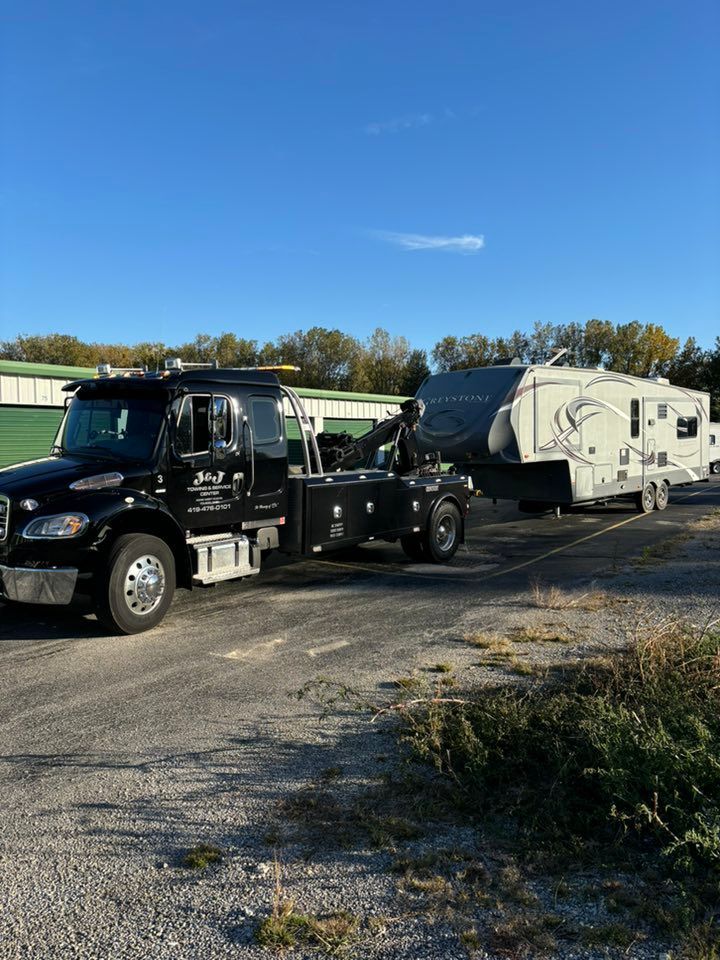 Black tow truck towing a white and gray fifth-wheel RV, parked on gravel in front of a green building.