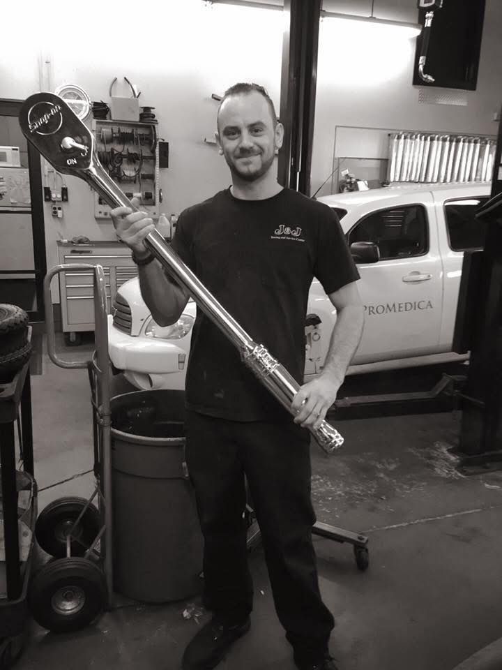 Mechanic holding a very large wrench in a garage, smiling. White truck in the background.