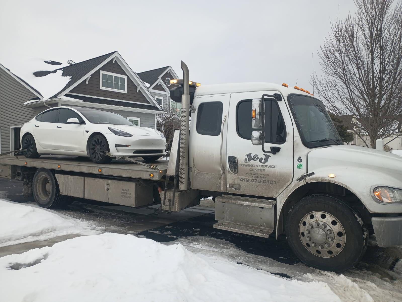 White Tesla on a tow truck in a snowy residential area. The truck is white and gray.