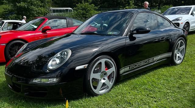 Black Porsche coupe on green grass with other cars in the background.
