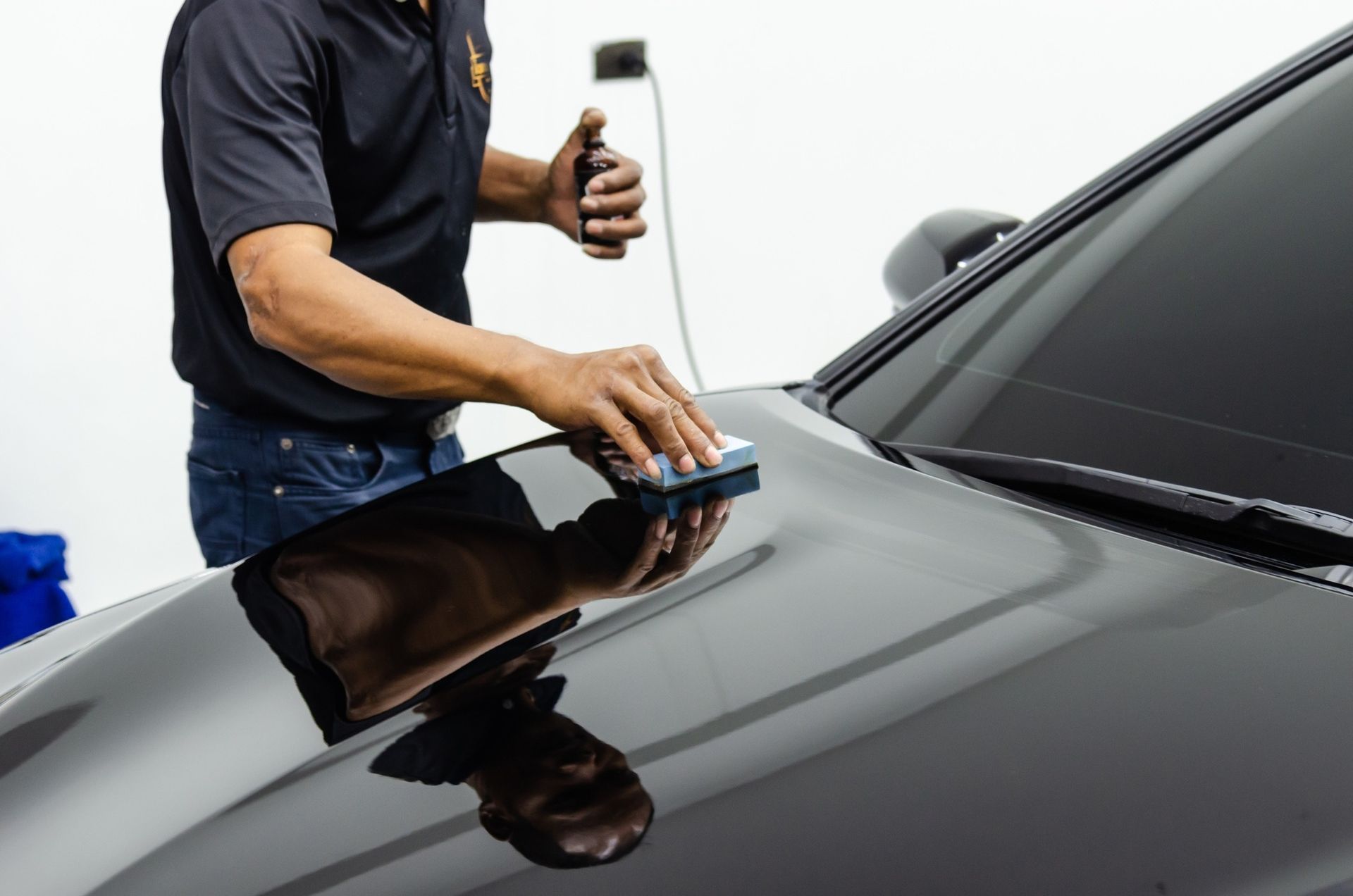 Person applying a coating to a black car's hood, using a sponge and bottle. White interior setting.