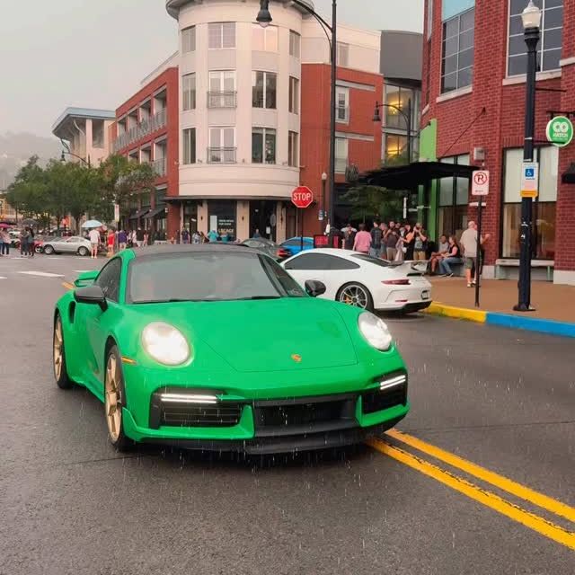 Green Porsche 911 Turbo S on a wet city street, gold wheels, white Porsche behind it, people in background.