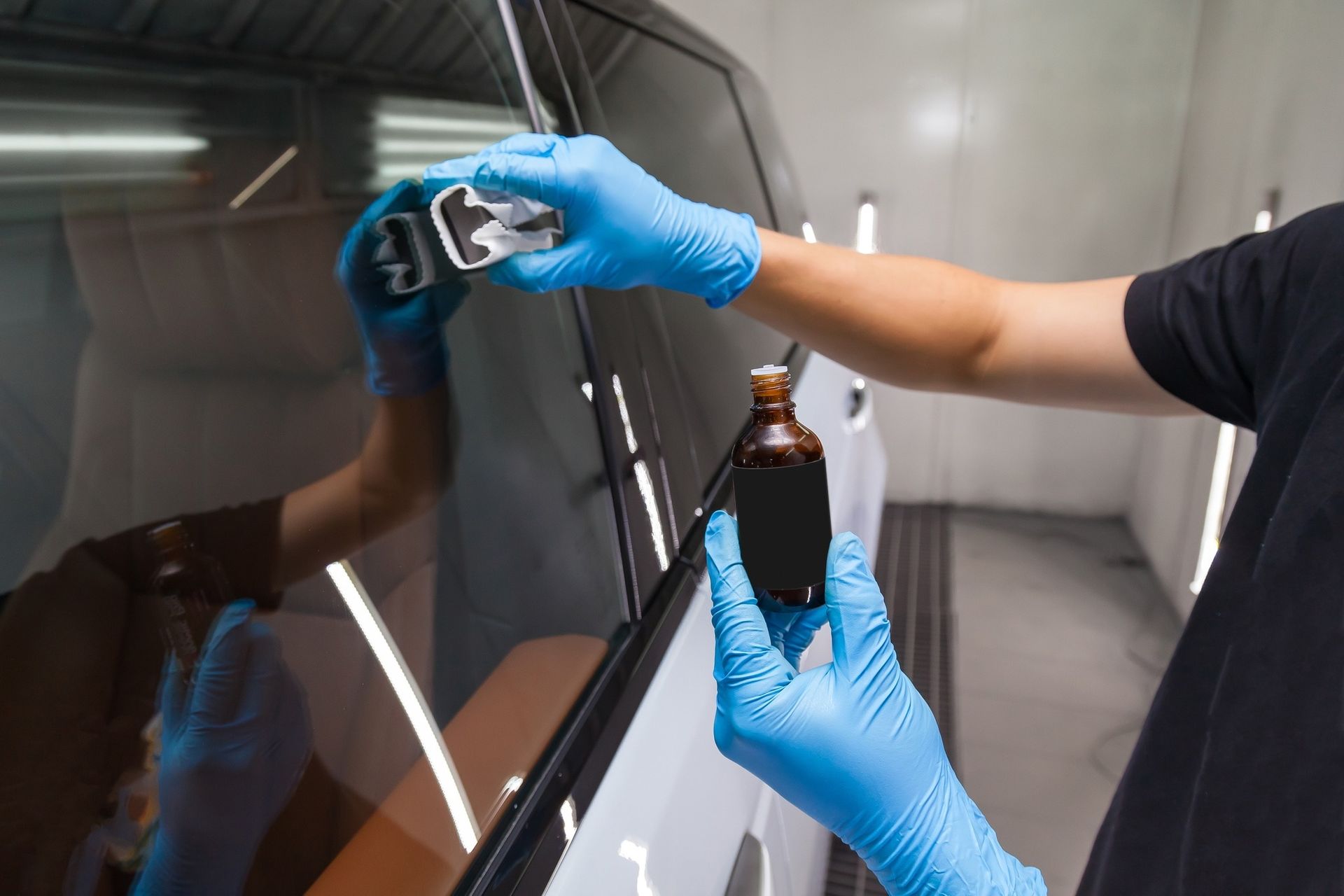 Person applying car window tint. Hands wearing blue gloves hold applicator and bottle, in a shop setting.