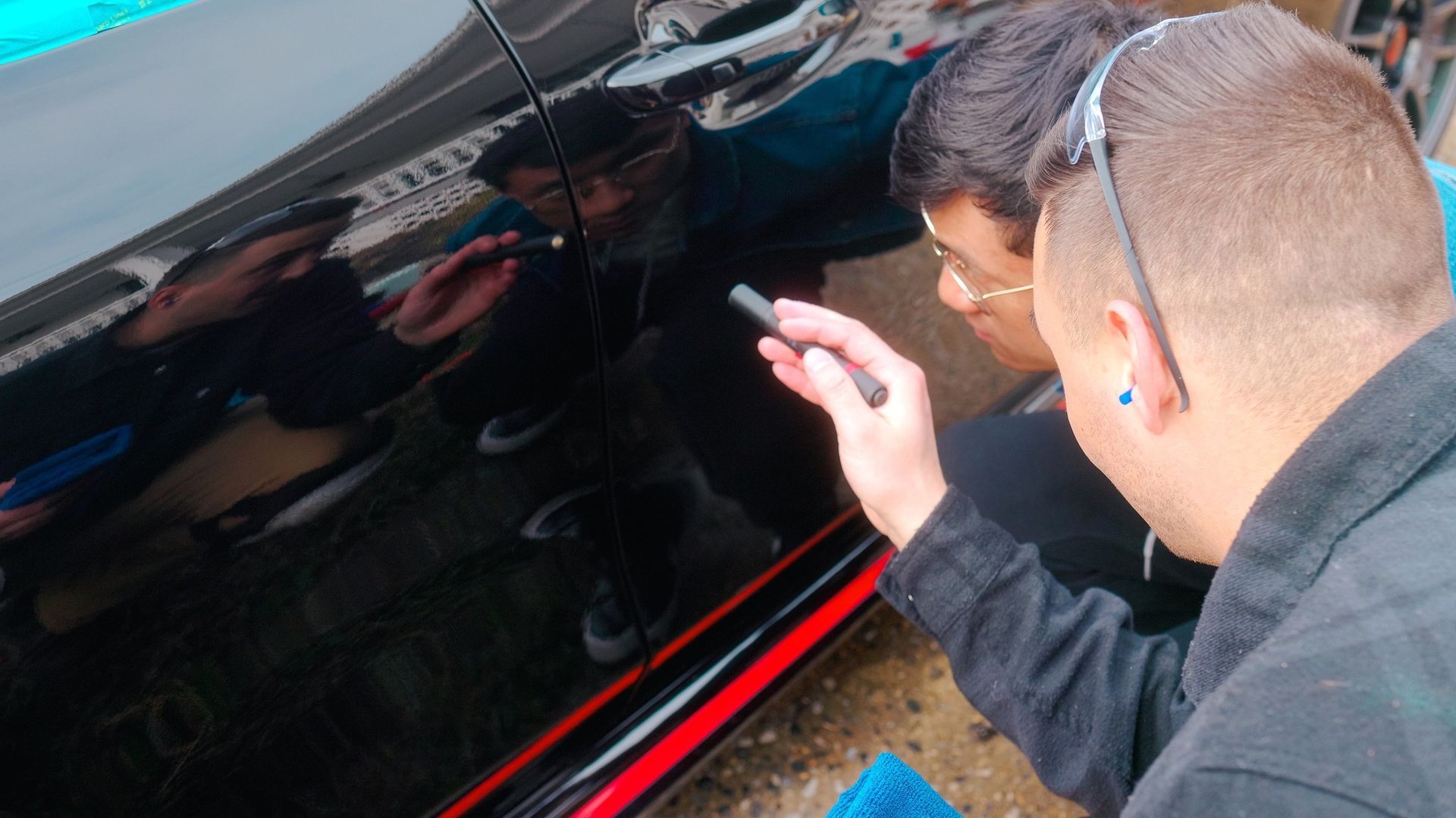 Two people inspecting a black car, one pointing with a pen. Bright reflections on the car's surface.