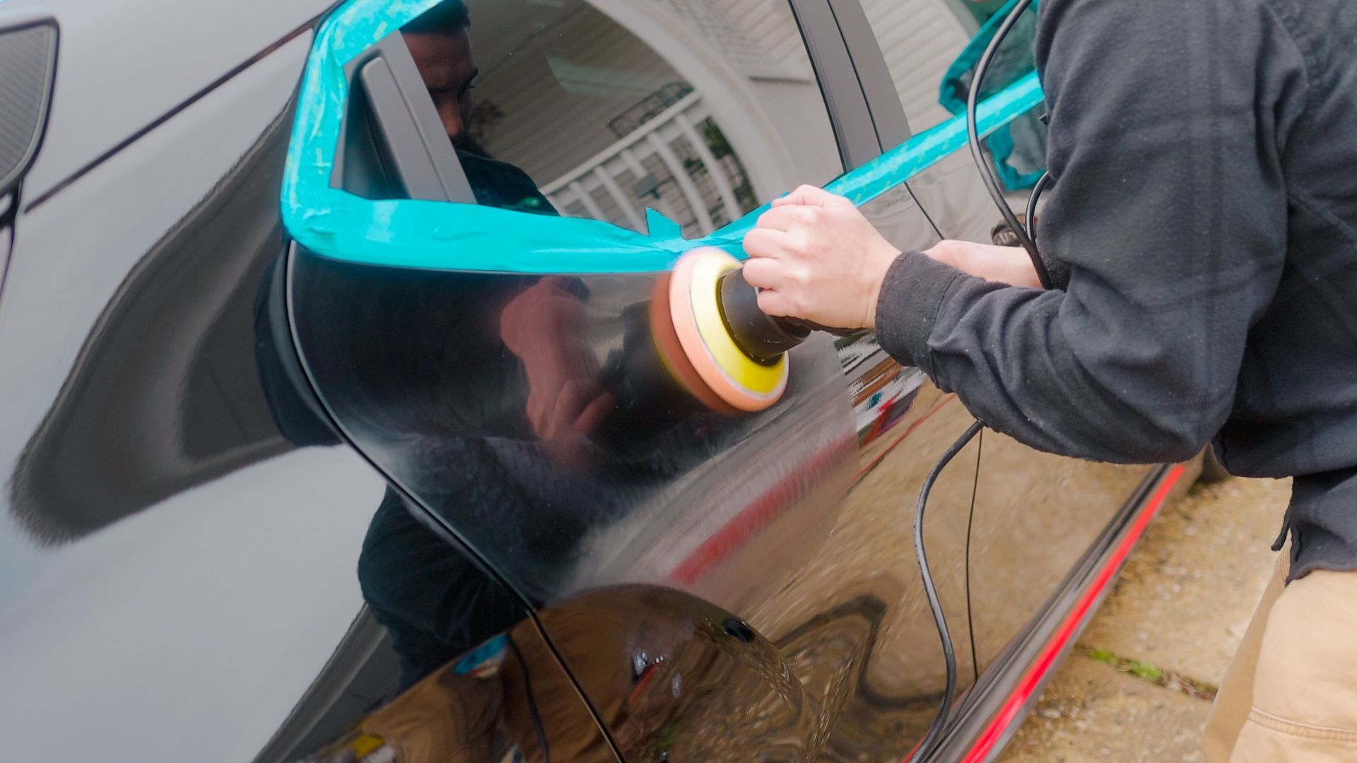 Person polishing a black car with a buffer; turquoise tape protects trim.
