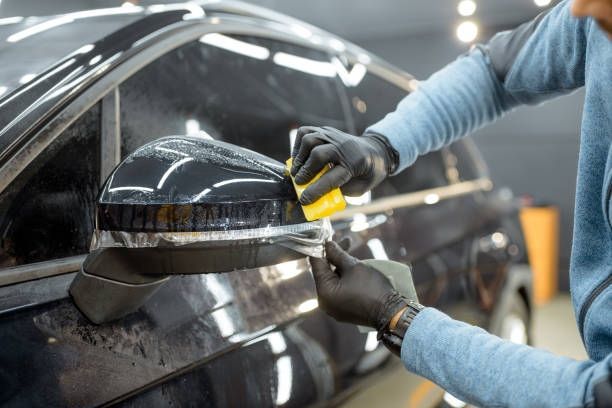 Person wearing black gloves applying film to a car's side mirror, in a workshop setting.