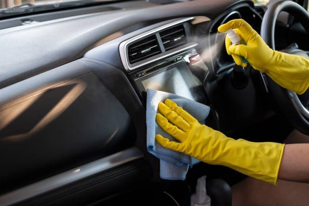 Hands in yellow gloves cleaning car dashboard with spray and cloth.