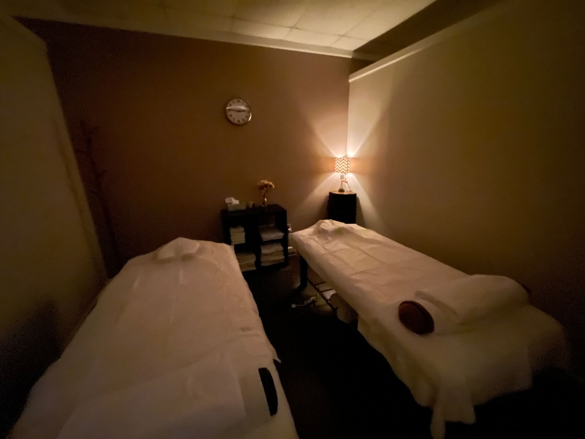 Two massage tables in a dim spa room with a clock and small table with items.