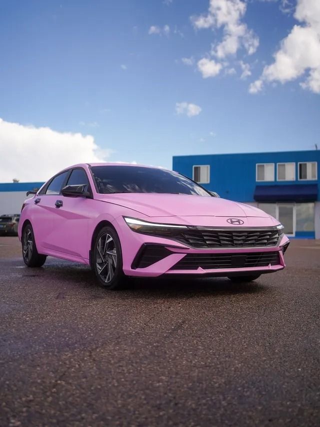Pink sedan parked on wet pavement beside a blue building under a cloudy sky