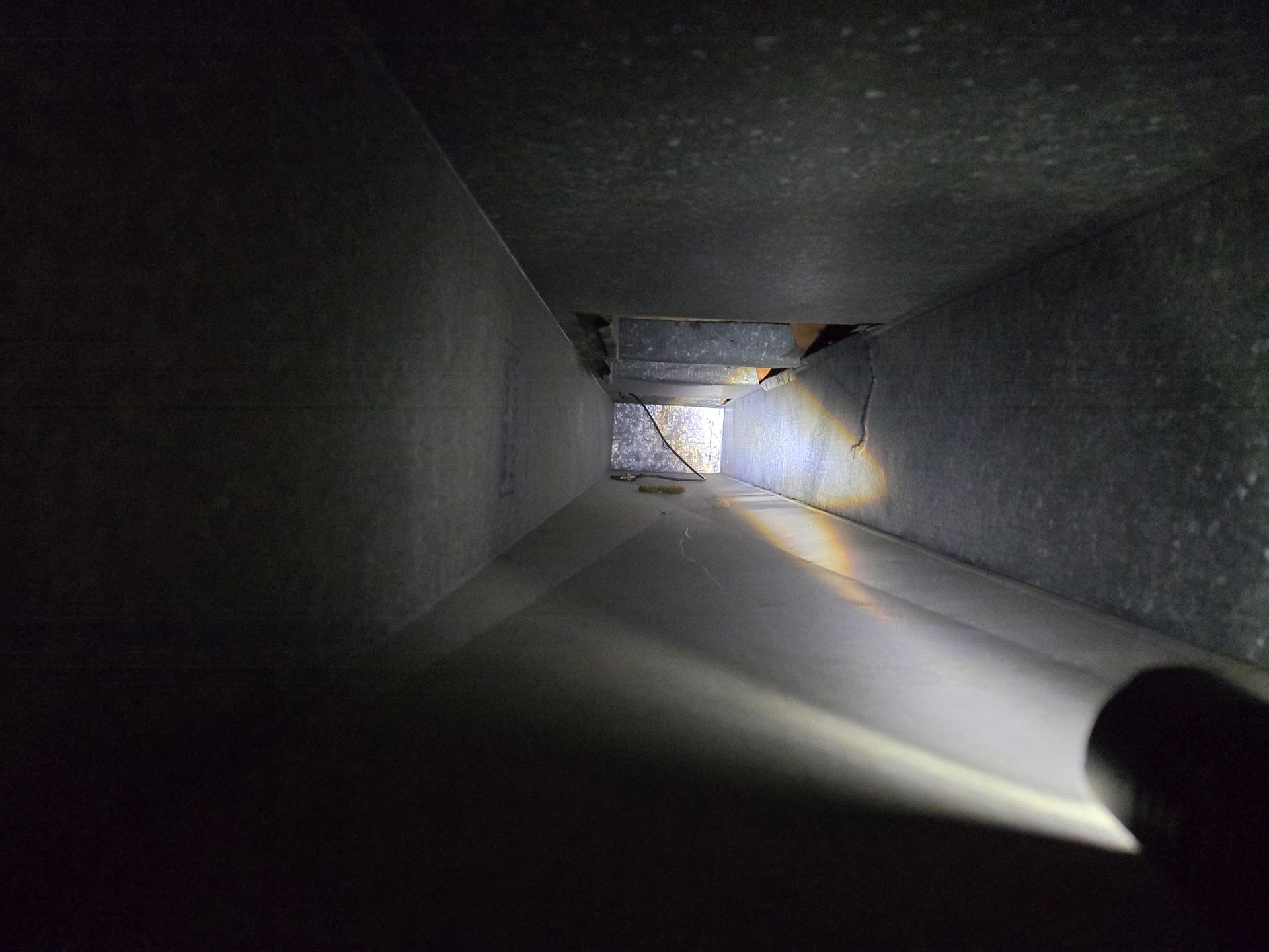 Inside a rectangular duct, illuminated by a flashlight. Gray metal walls with some debris visible.
