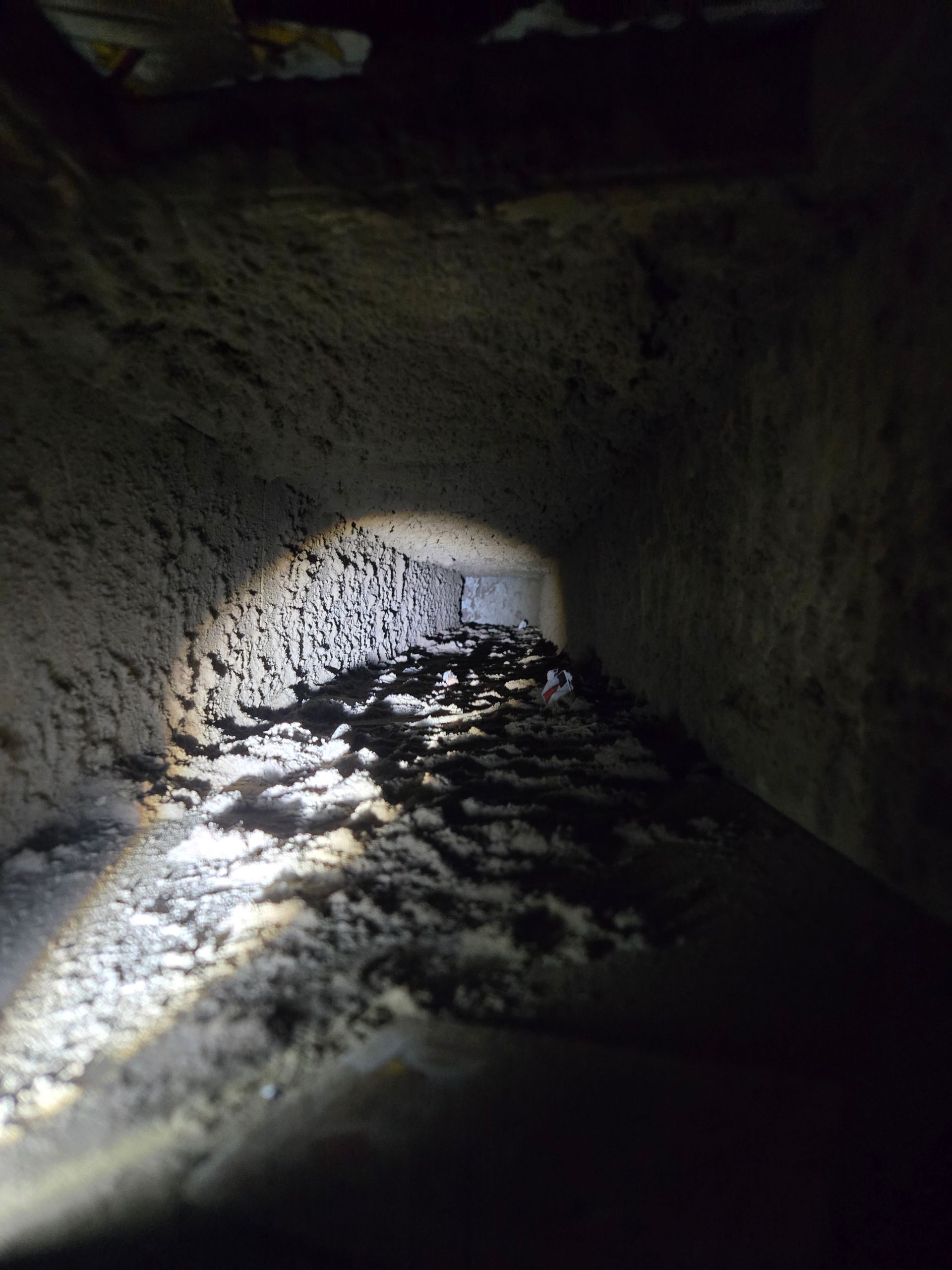 Inside of a dark, dusty air duct illuminated by a flashlight.