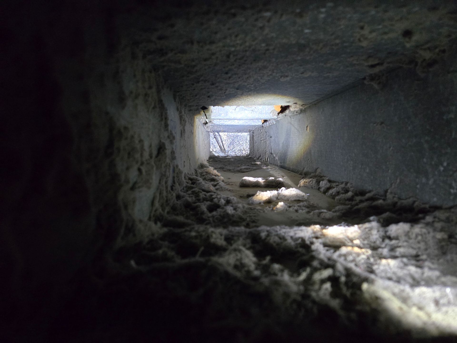 Inside view of a dusty air duct with a light at the end, showing layers of debris.
