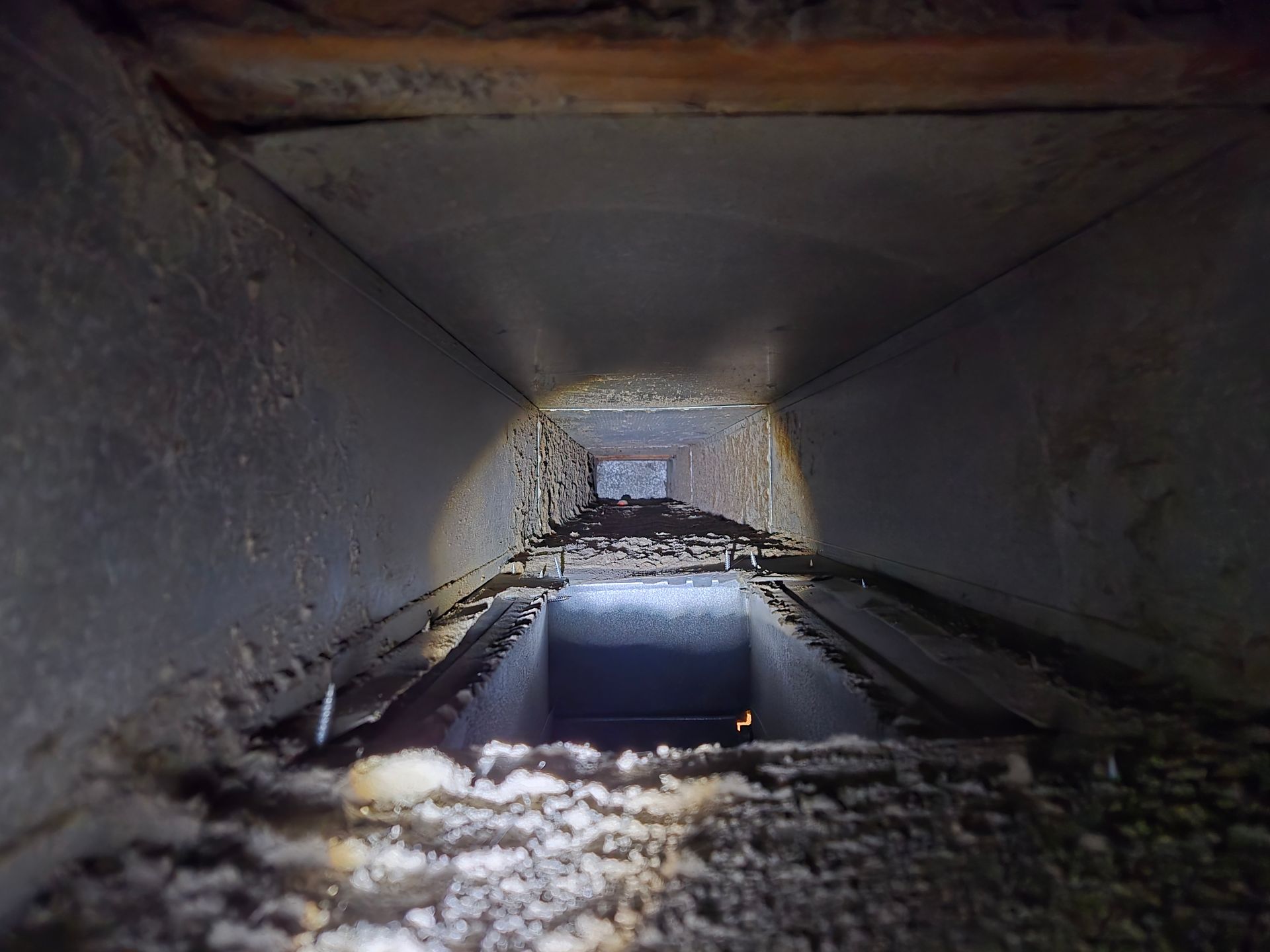 Inside of a dusty HVAC duct. The walls are covered in dust and debris, with a dark opening at the end.