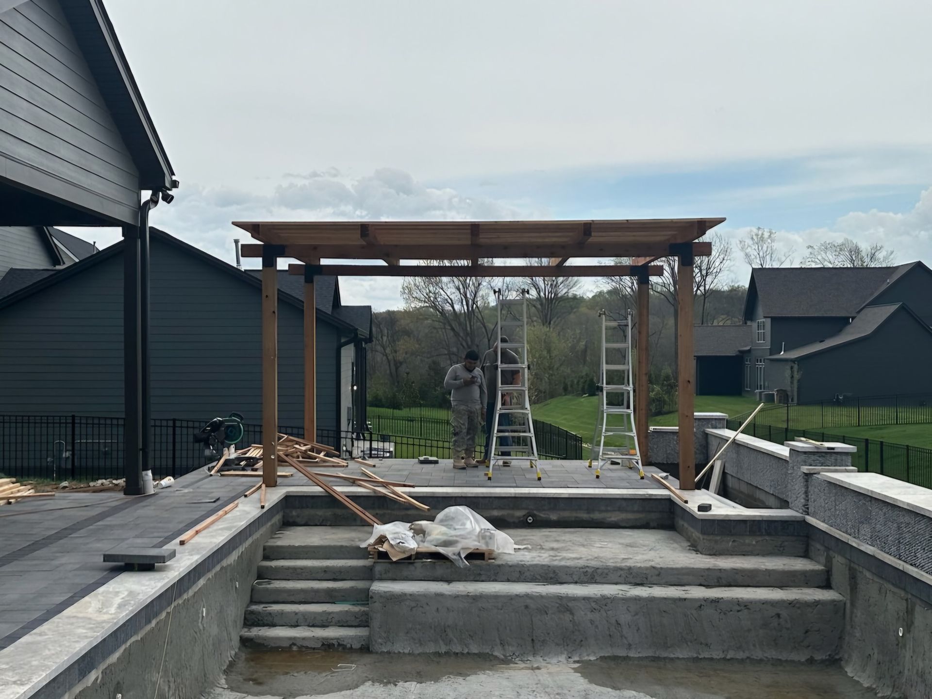Backyard patio under construction with wooden pergola, concrete steps, and a ladder in the center
