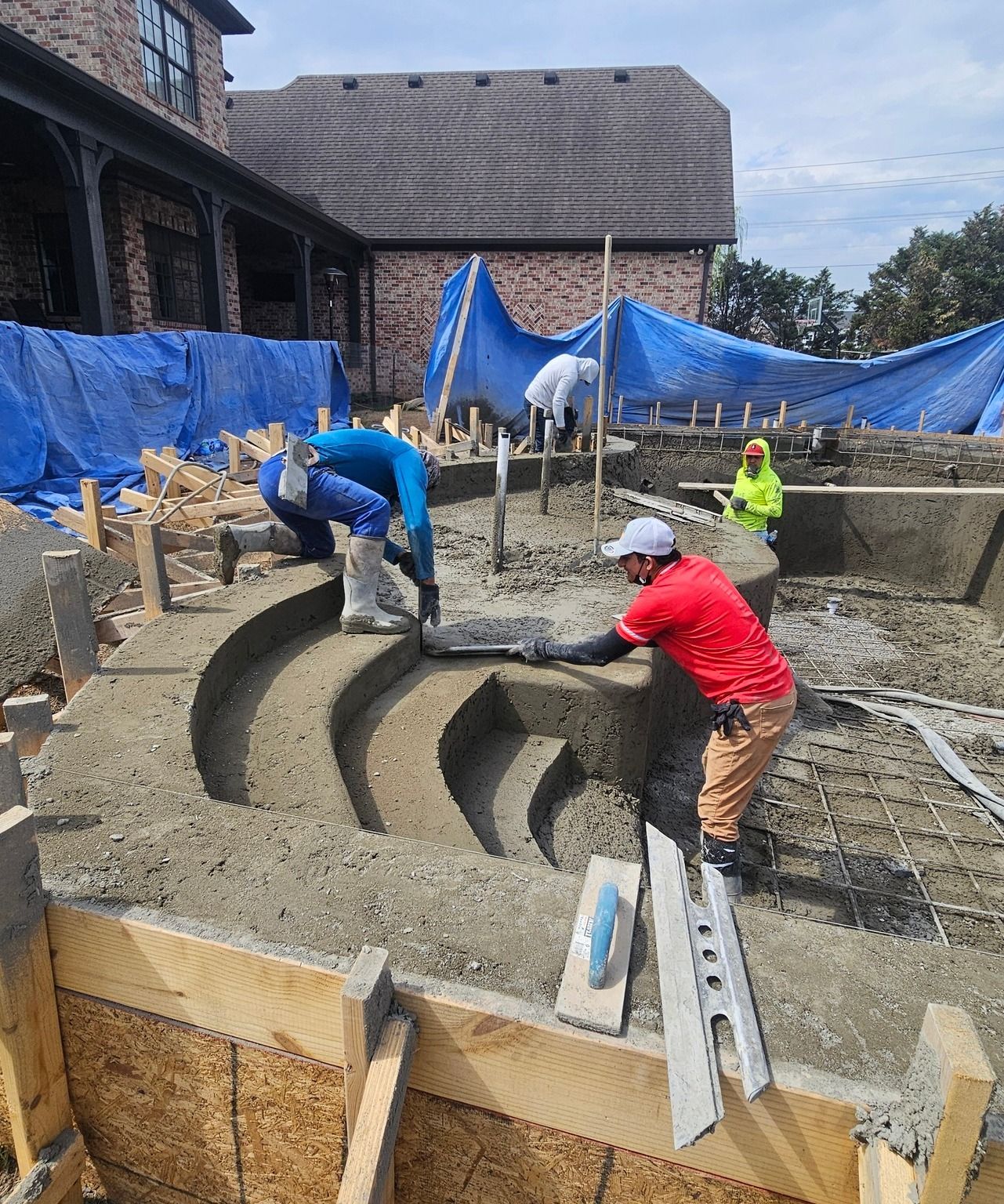 Workers shaping a curved concrete pool in a backyard construction site with blue tarps and a house nearby