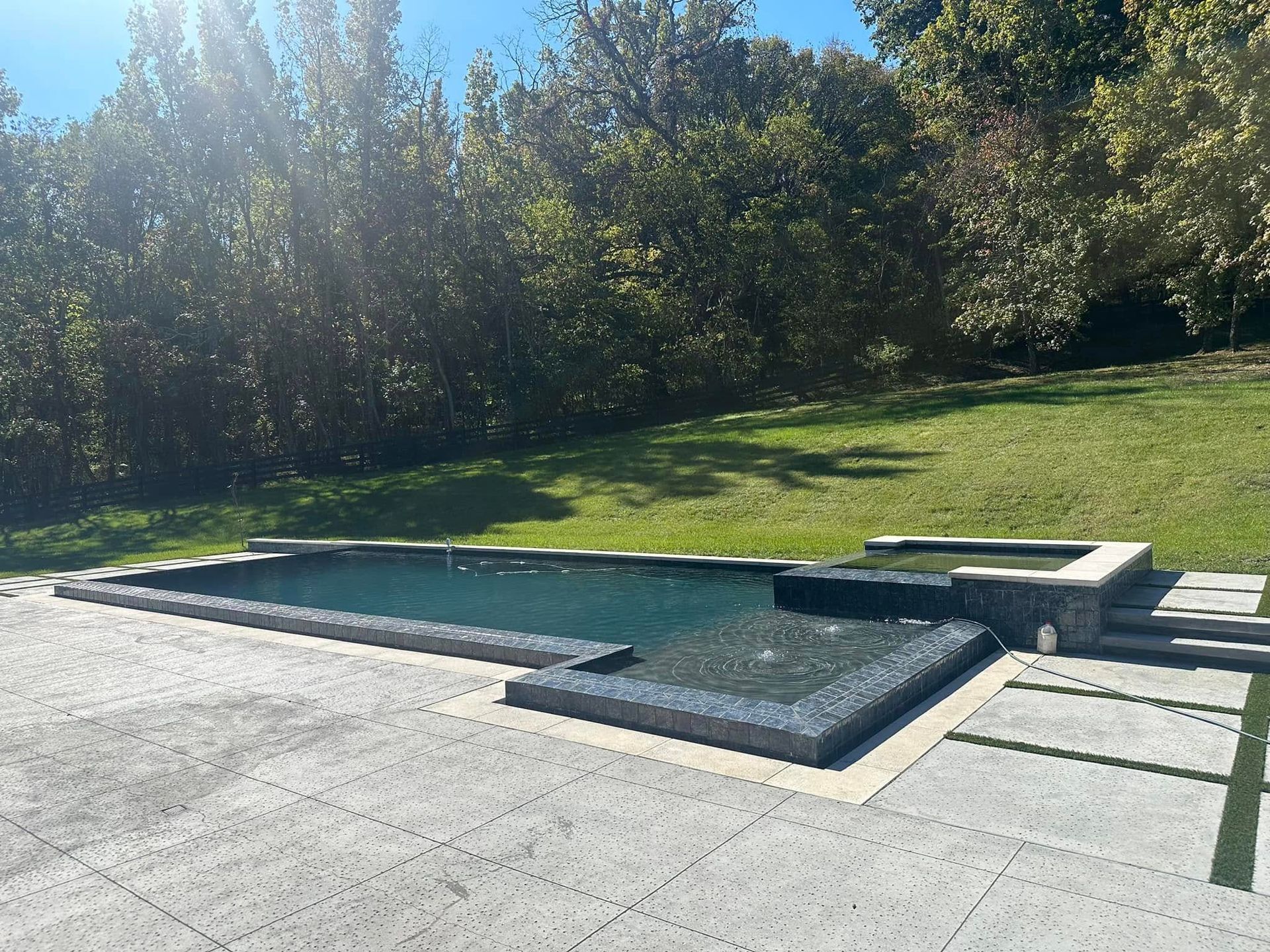 Outdoor pool with two blue spa basins beside a concrete deck and grassy trees in the background