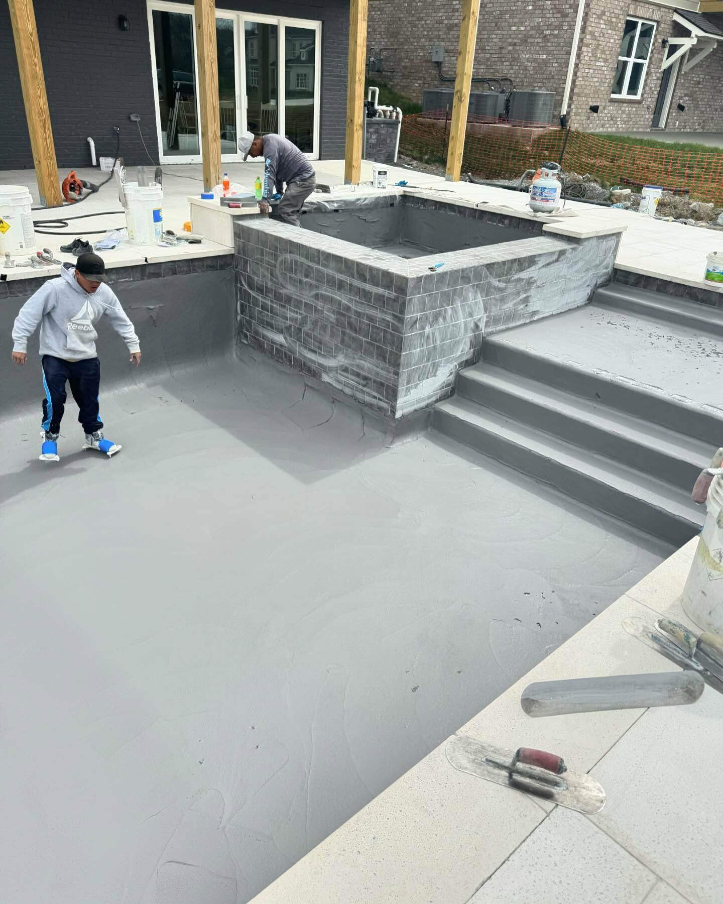 Workers finishing a gray concrete patio and steps beside a house, with tools on the surface