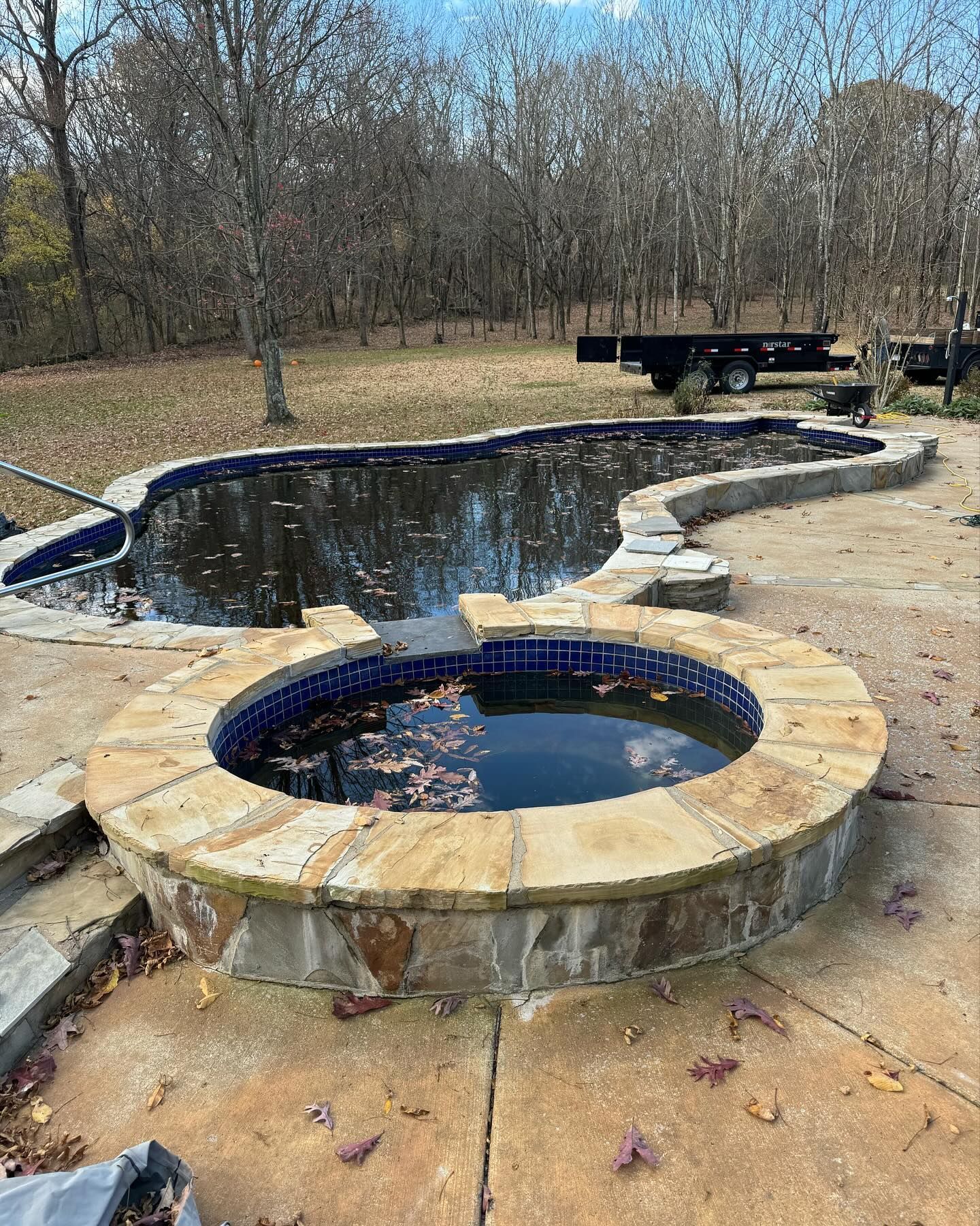 Partially filled stone pool and circular hot tub on a concrete patio in a wooded backyard