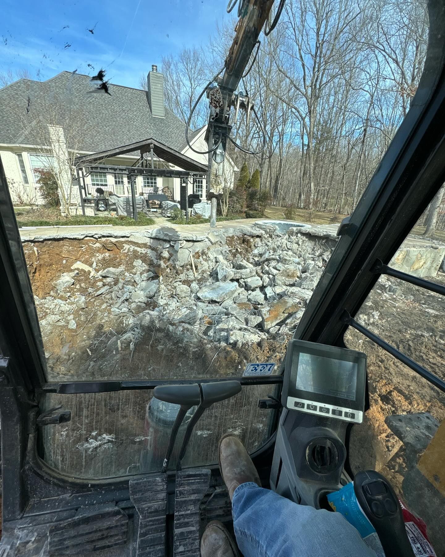 Inside a construction cab, a machine is pushing dirt and rocks beside a house in winter.