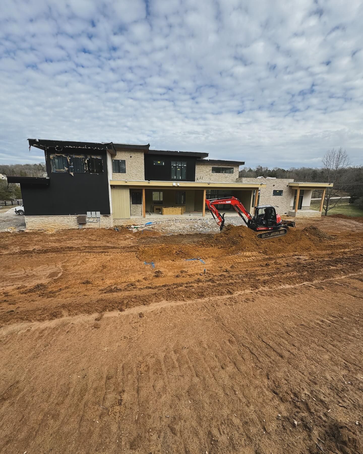 House under construction with a red excavator in front and a cloudy sky overhead