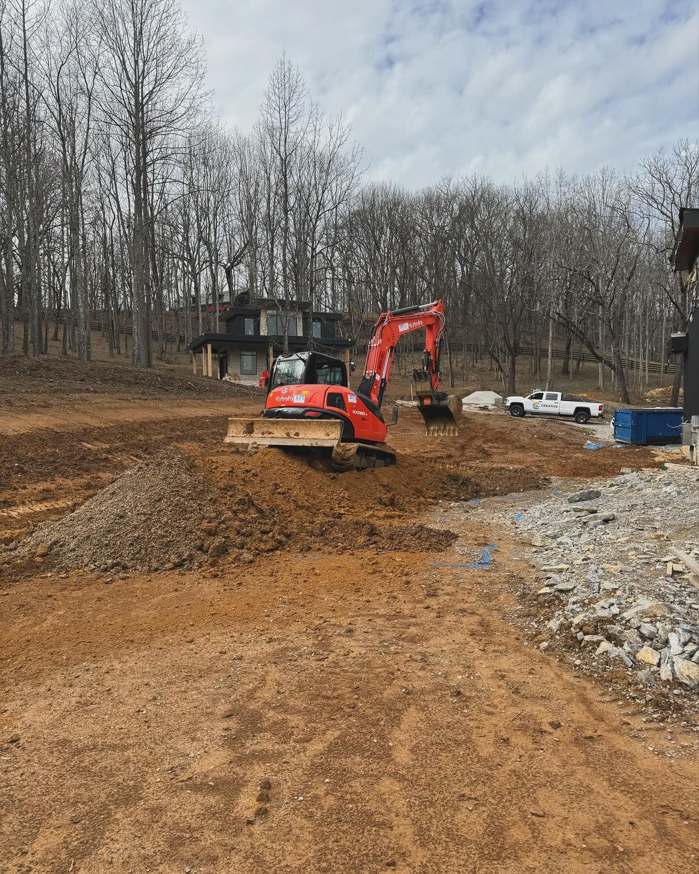 Red excavator on a dirt lot with gravel piles, trees, and a small cabin in the background