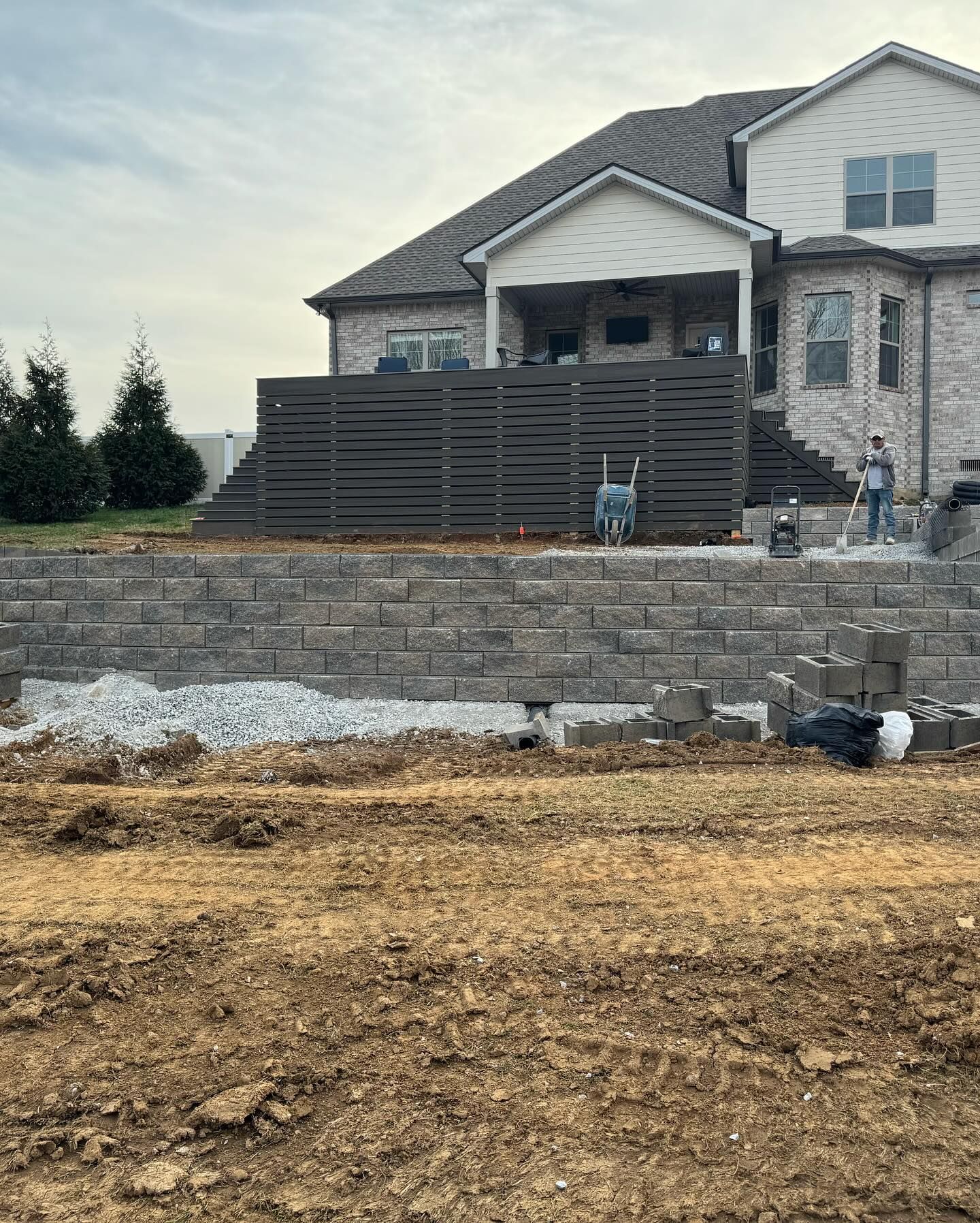 Stone house under construction with a retaining wall and workers on a dirt lot under overcast skies