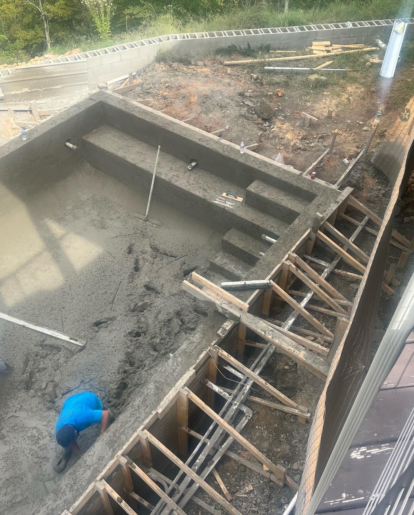 Worker in blue shirt inside a concrete foundation pit with wooden bracing and muddy ground.