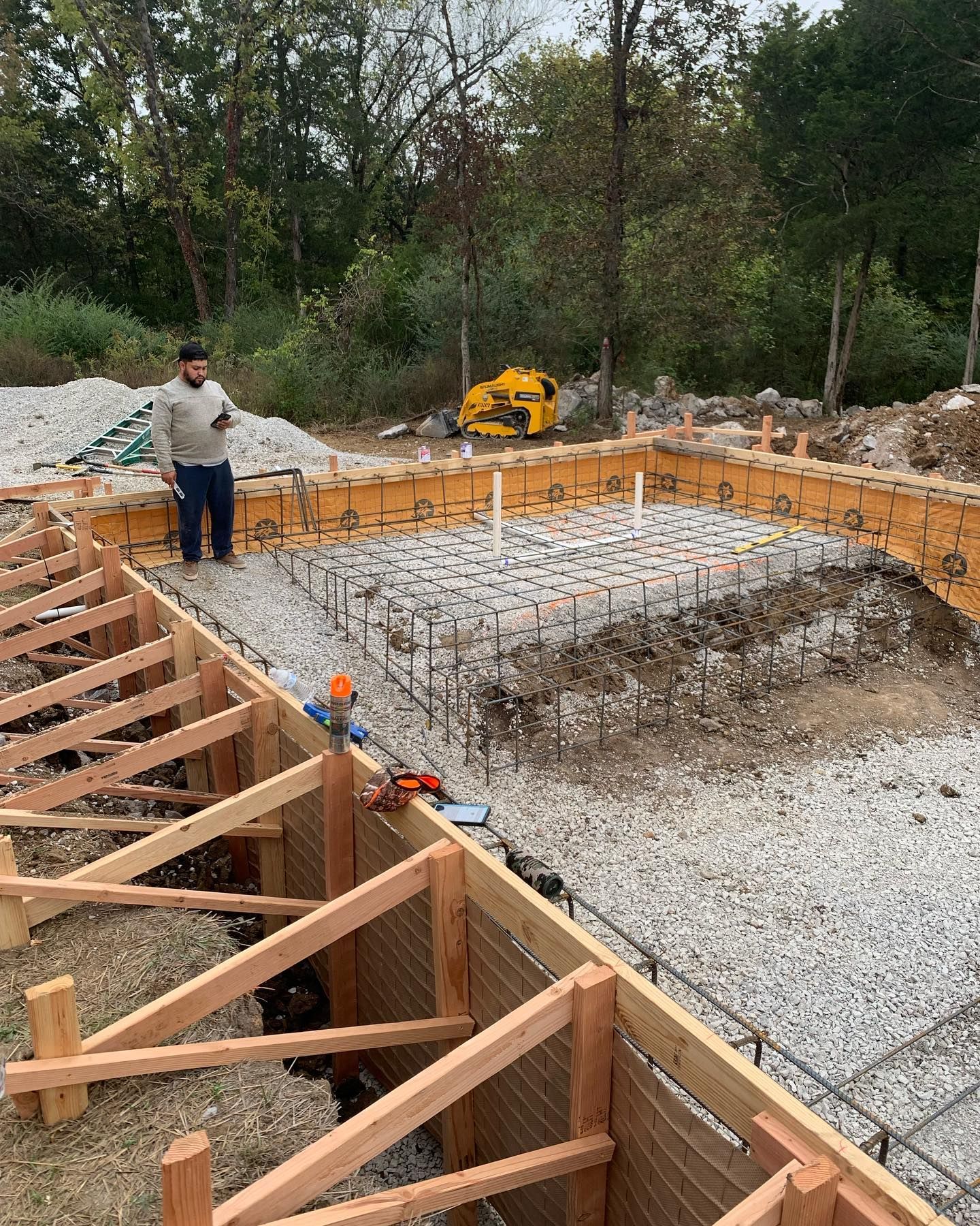 Foundation construction site with wooden formwork, rebar grid, gravel, and a worker near piles of stone
