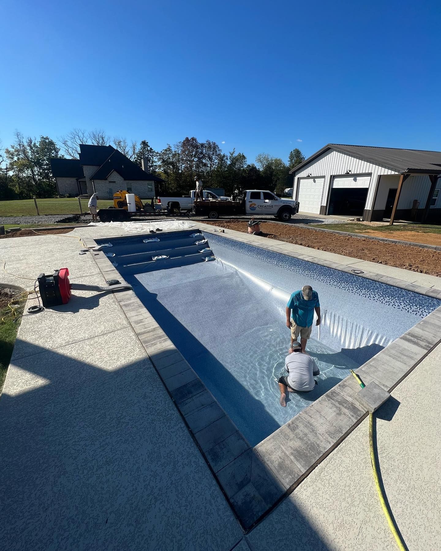 Workers coating an empty backyard pool with white liner material beside a house and garage.