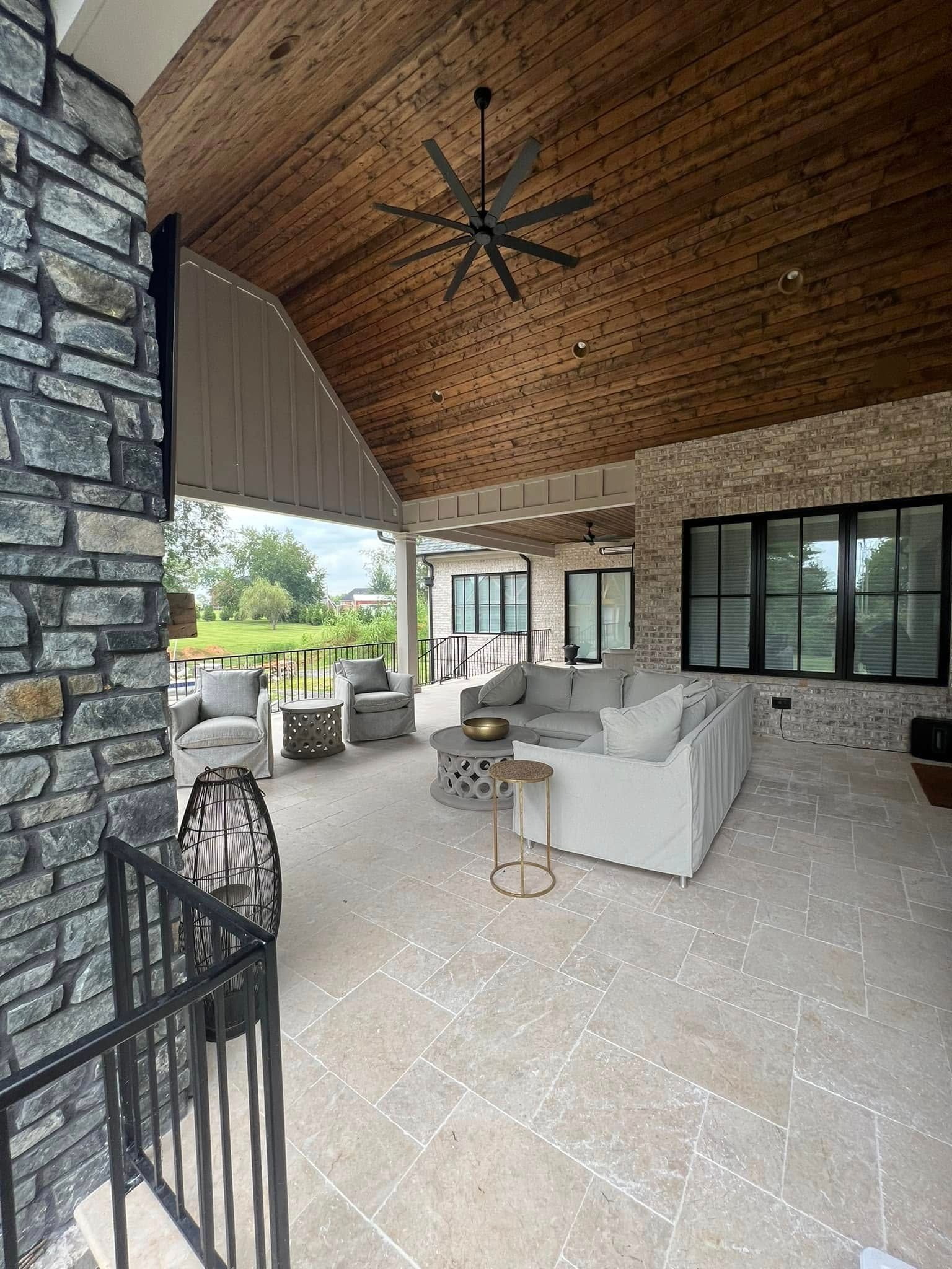 Covered patio with stone fireplace, wood ceiling, white seating, and large windows overlooking a yard