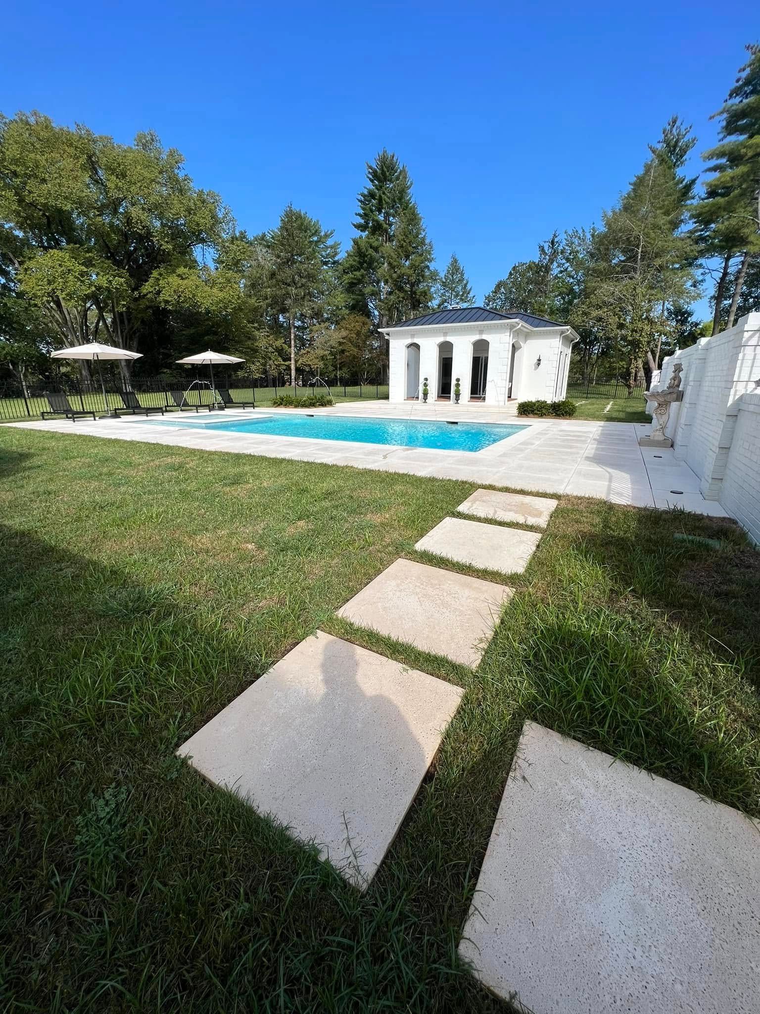 Backyard pool with white cabana, stepping stones, green lawn, and trees under a blue sky