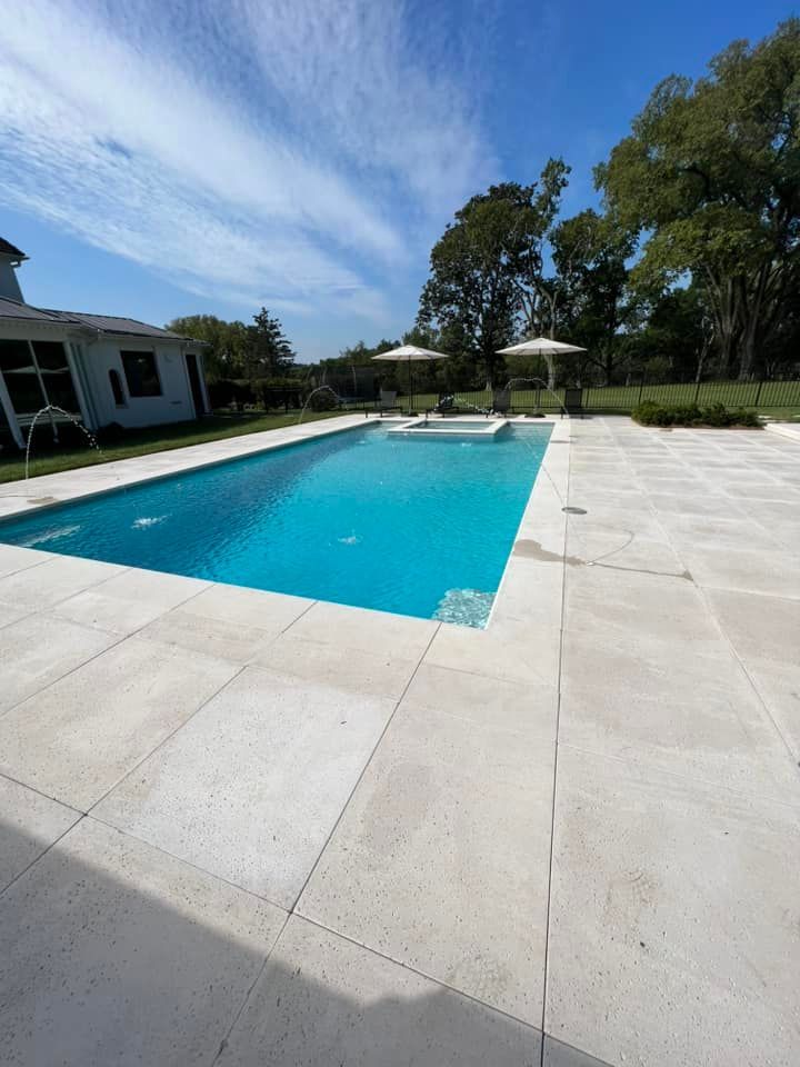 Outdoor swimming pool with turquoise water, white stone deck, and trees under a blue sky