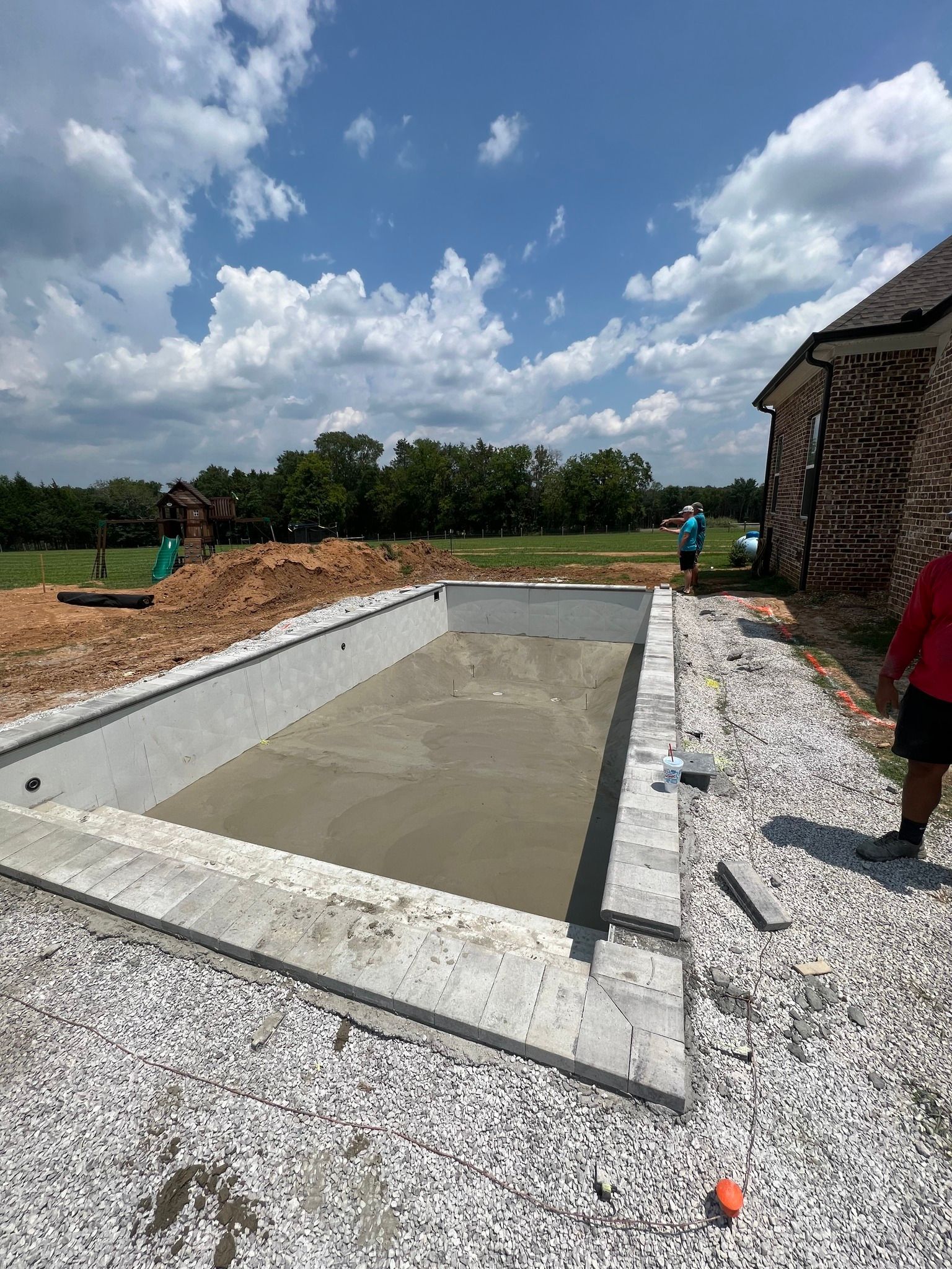Concrete pool or foundation under construction beside a brick building on a gravel site.