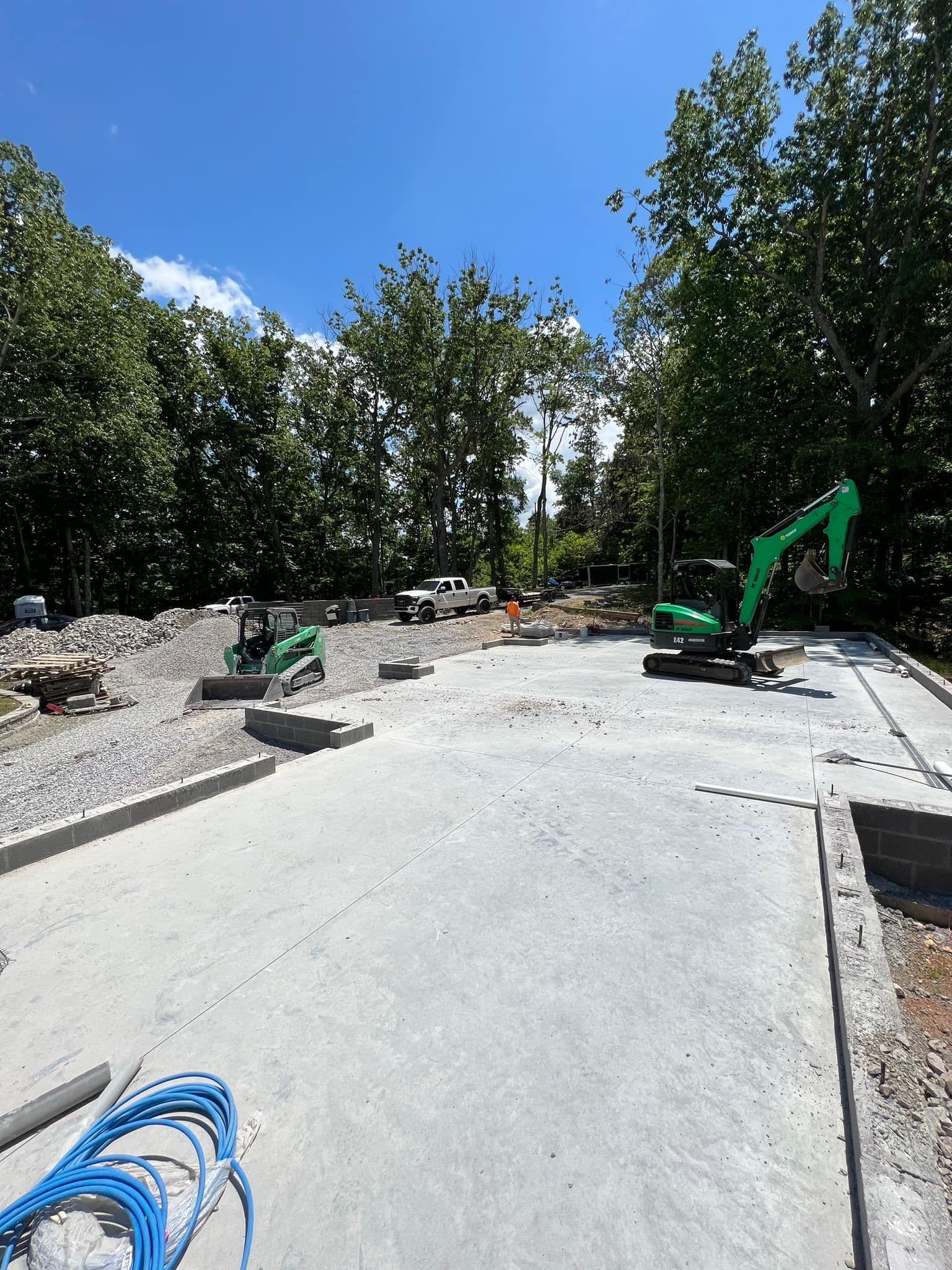 Construction site with freshly poured concrete slab, excavator, and trees under a clear blue sky