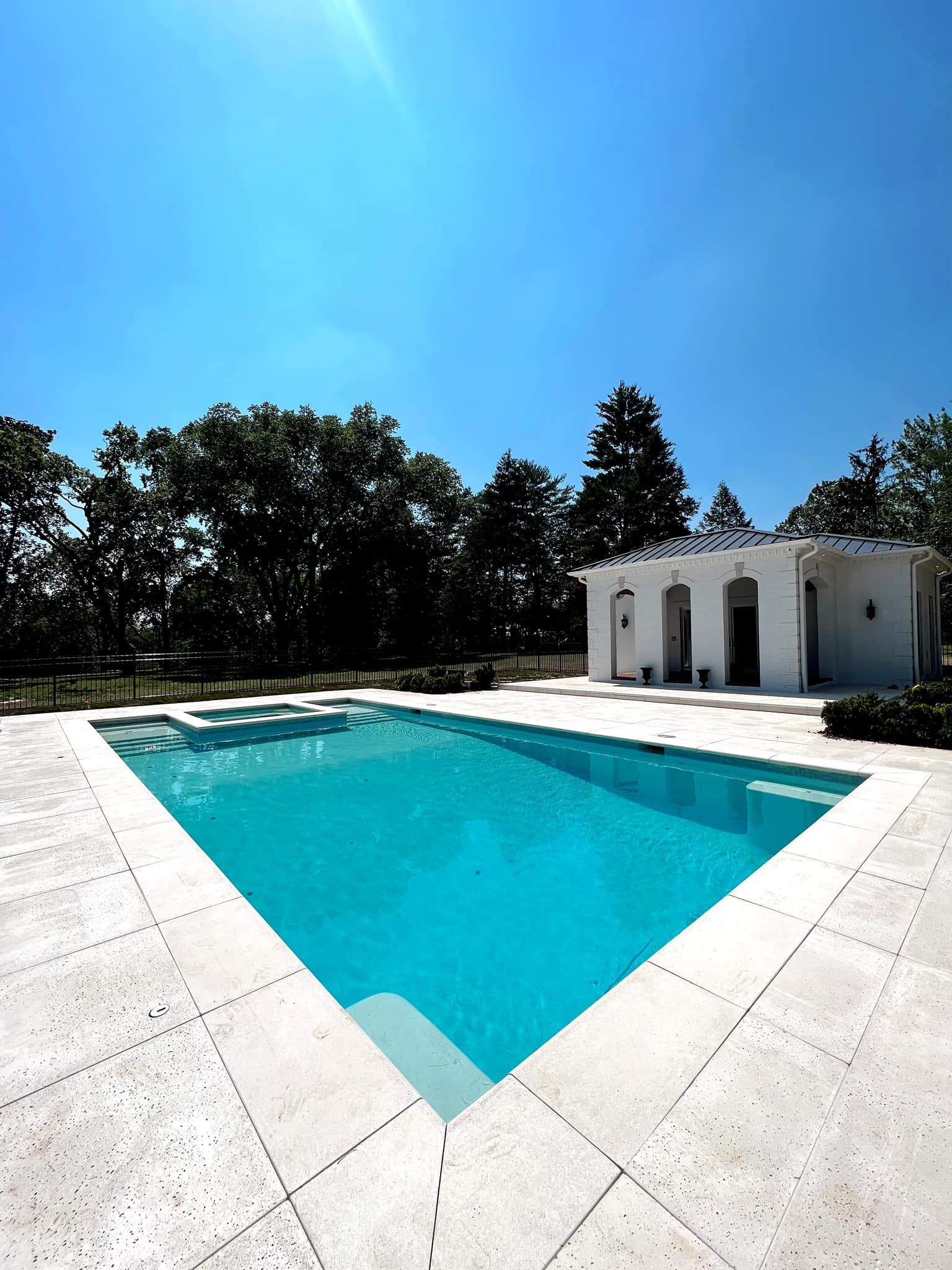 Bright turquoise outdoor pool beside white stone patio and a small white building under a clear blue sky