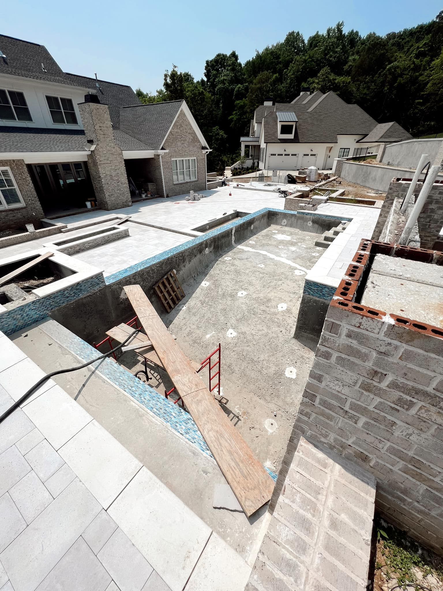 Backyard pool under construction beside a stone house, with white tile, concrete, and scaffolding.