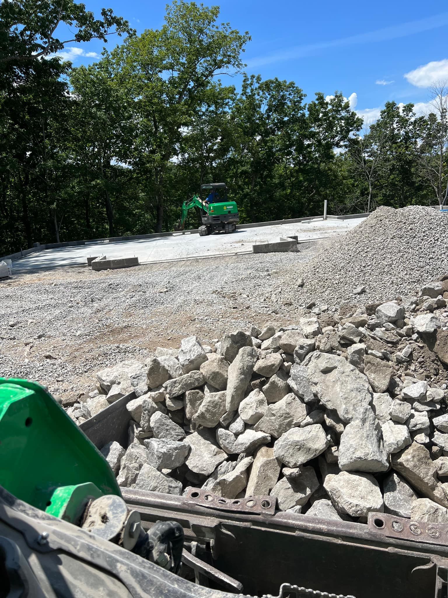 Rocky construction site with piles of gravel, a green machine, and trees under a blue sky