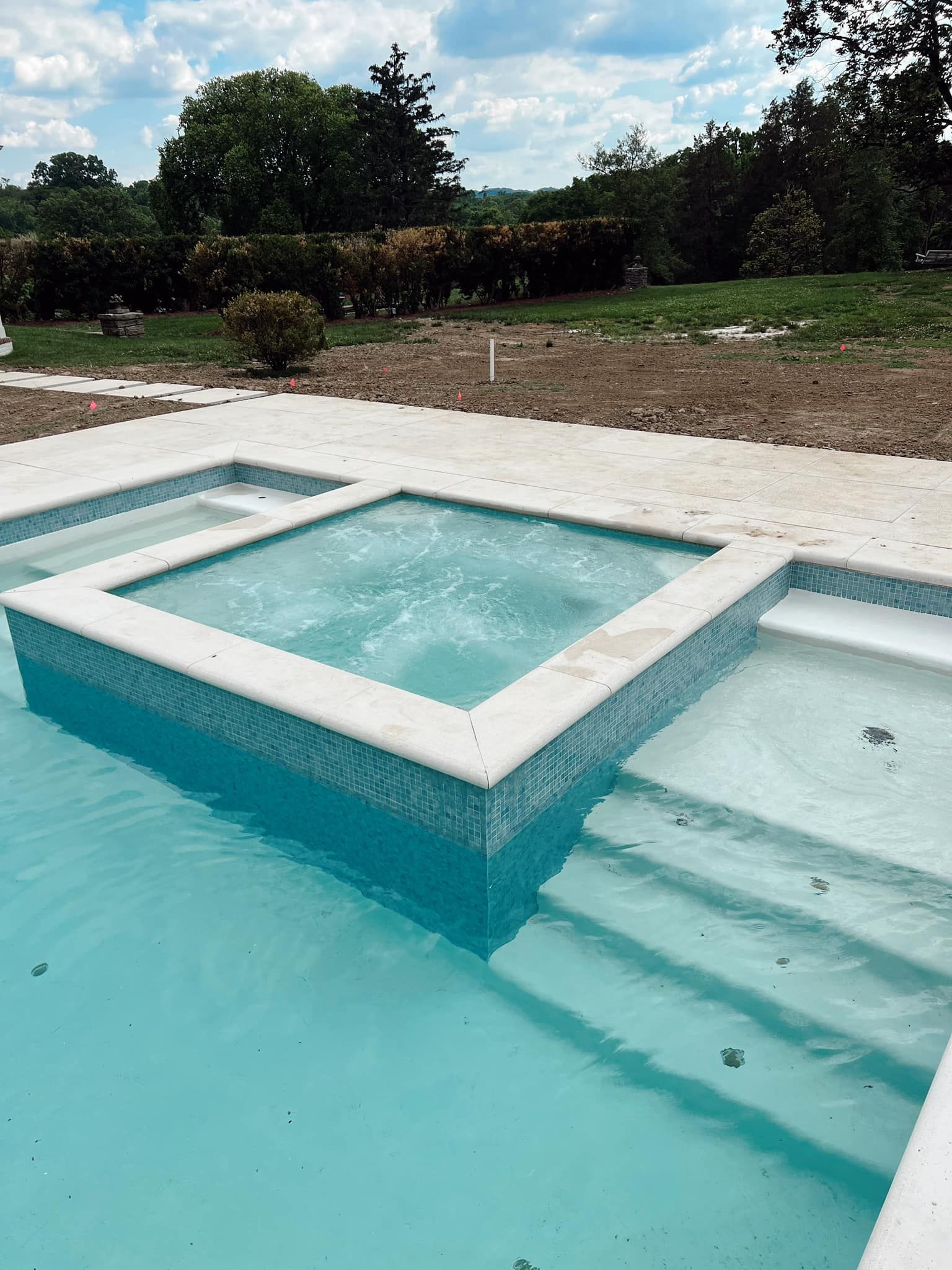 A turquoise pool with a raised rectangular spa and steps, set beside a gravel yard and trees.